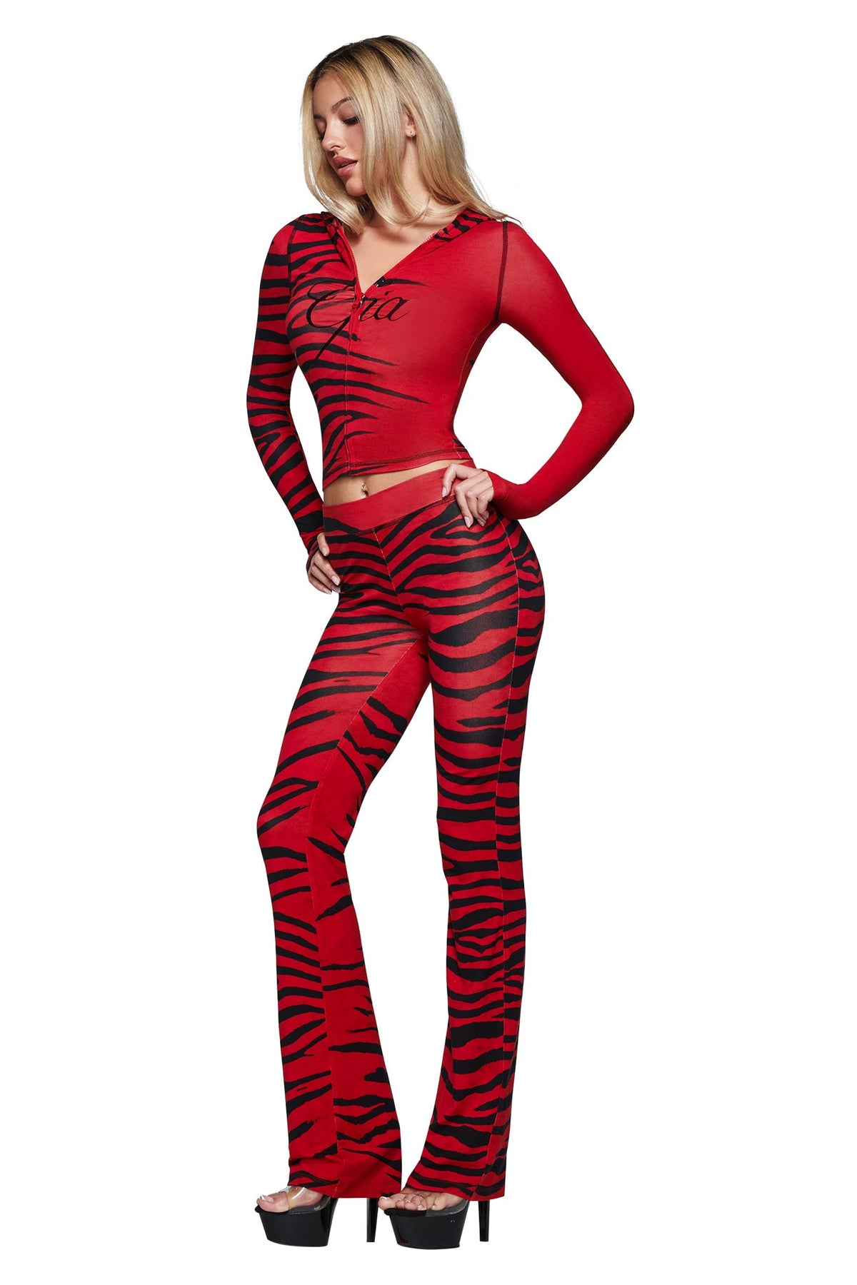 A woman models the I.AM.GIA BLARE HOODIE - RED WILD SIDE, a red and black striped two-piece with long sleeves and flared pants, crafted from soft bamboo fabric, paired with black platform heels against a white background.