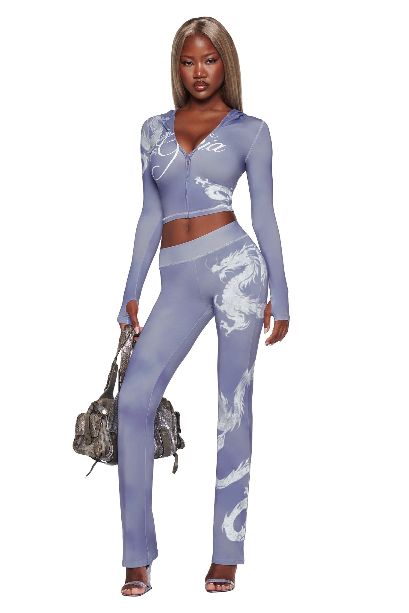 A woman wearing the I.AM.GIA BLARE HOODIE—STEEL BLUE DRAGON, featuring bold white dragon motifs, paired with a fitted blue look, silver handbag, and high-heeled sandals.
