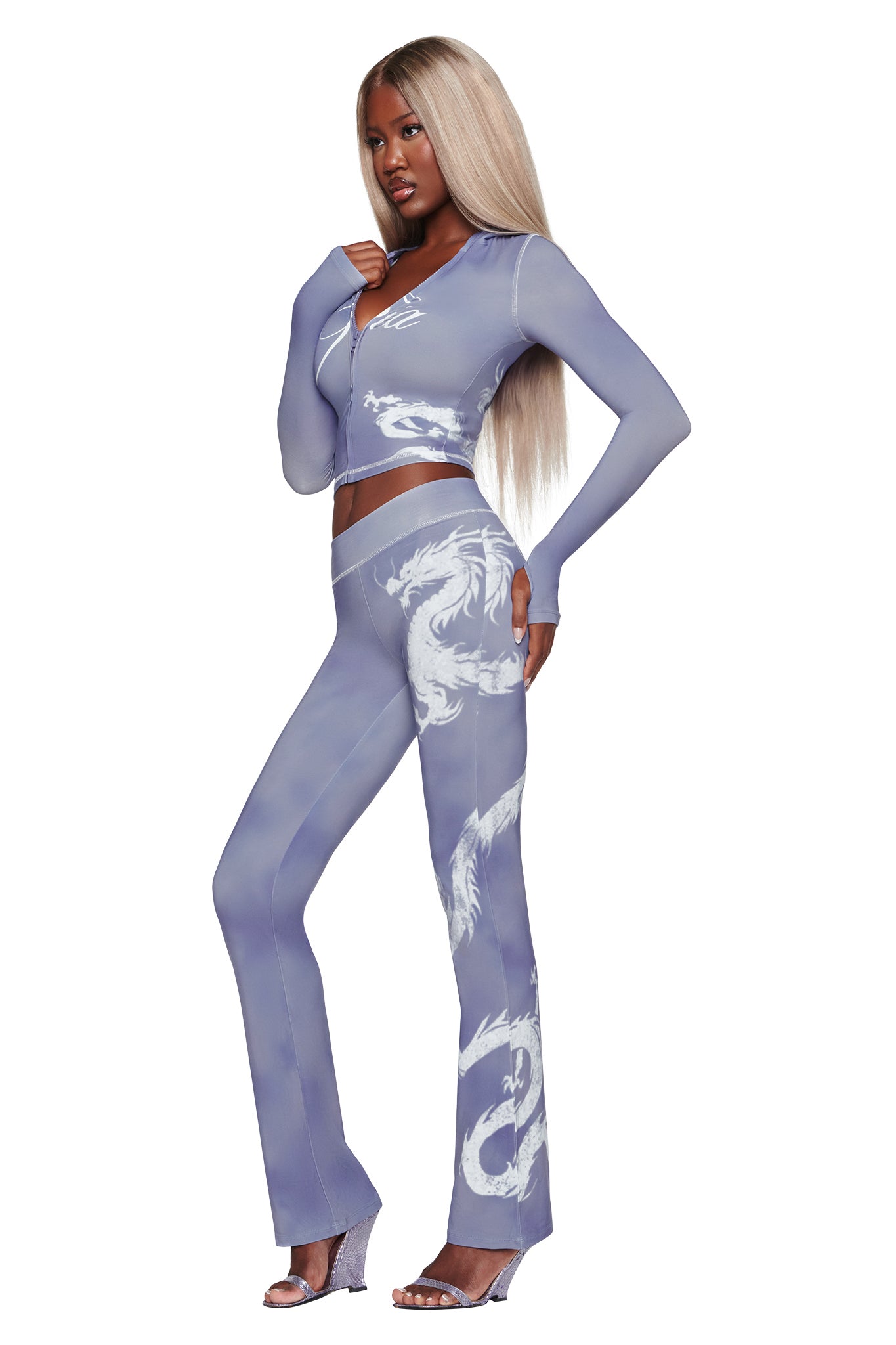 A woman with long blonde hair wears the BLARE HOODIE - STEEL BLUE DRAGON by I.AM.GIA, featuring fitted, sheer purple fabric adorned with white dragon designs inspired by the mystique of the Steel Blue Dragon. She completes the look with open-toe heels and poses confidently against a white background.