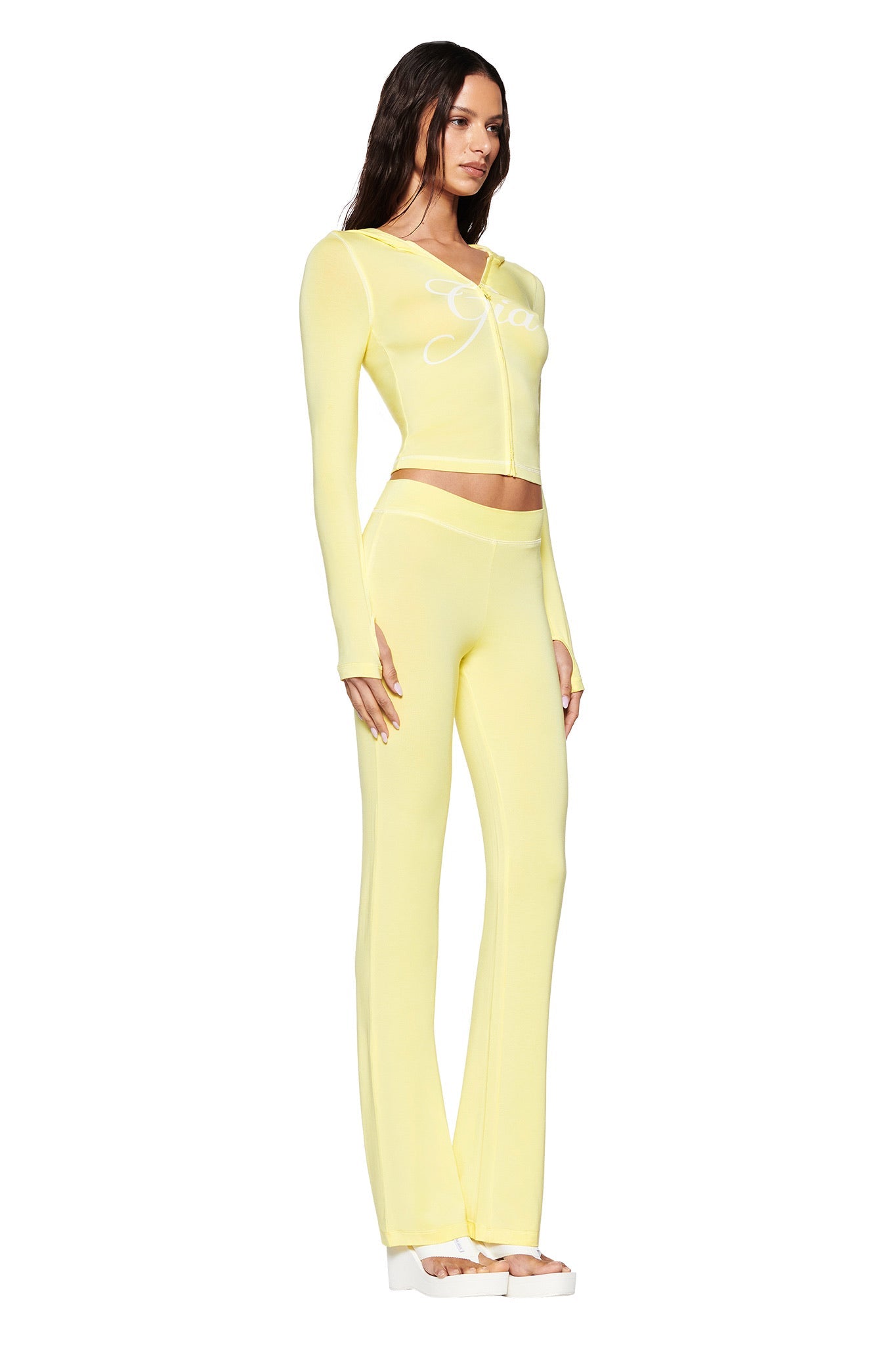 A woman models a vivid yellow outfit, showcasing a bodycon long-sleeve crop top and high-waisted pants. Her top features white text reminiscent of the I.AM.GIA Blare Hoodie. She pairs this with white platform sandals, all set against a white backdrop.
