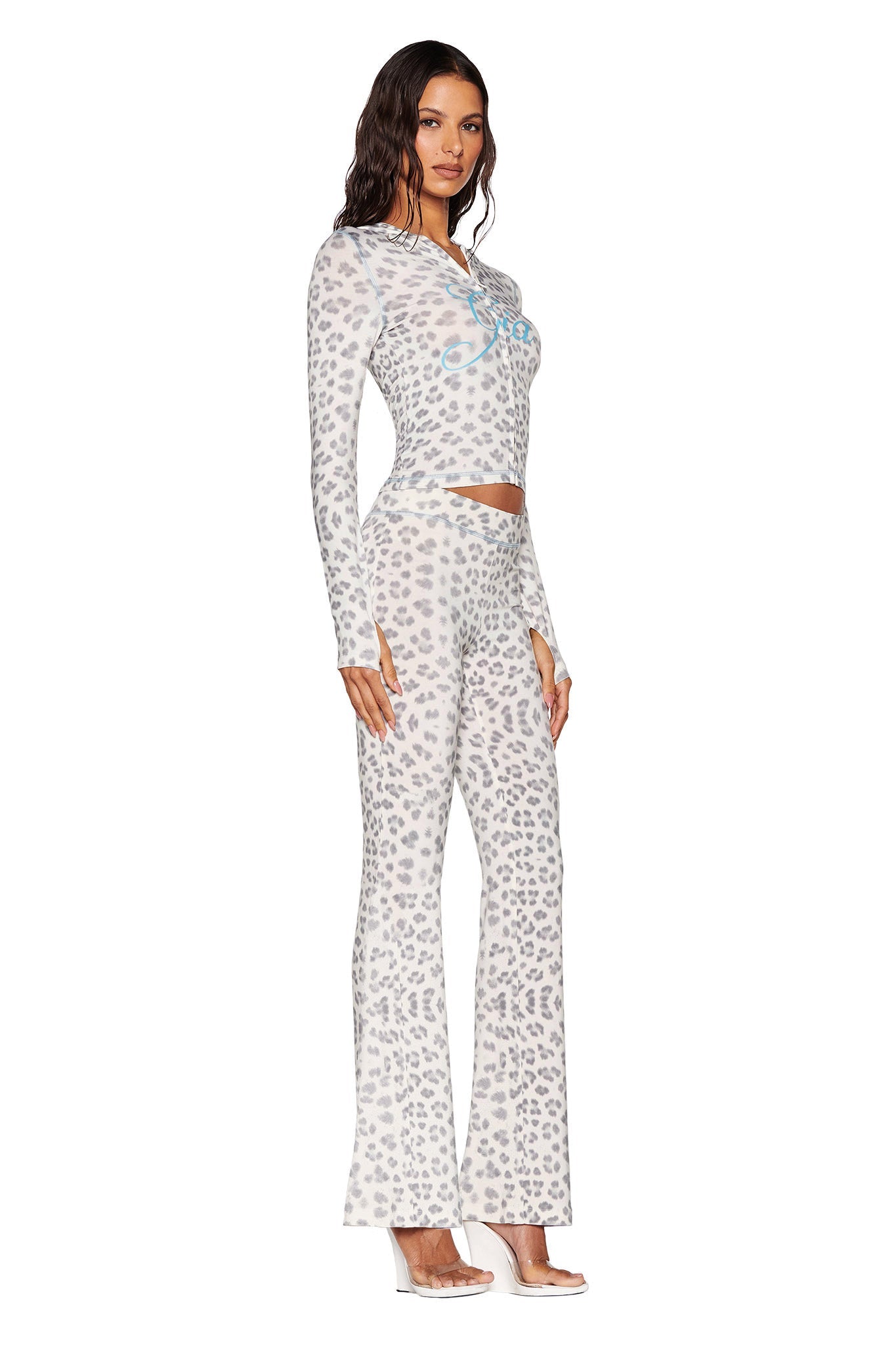 A woman confidently poses sideways against a plain white background, showcasing the BLARE TRACKPANT - SNOW LEOPARD : WINTER WILD CARD from I.AM.GIA. She wears this fitted, long-sleeve ensemble with clear high heels, her long, wavy hair adding to her striking look.