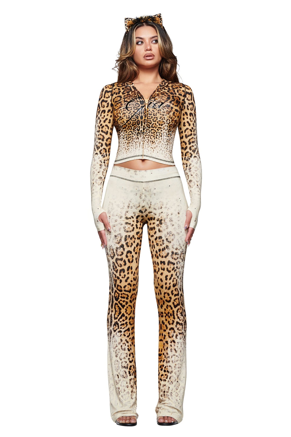 A woman stands against a white background wearing I.AM.GIA’s BLARE TRACKPANT in Cream Faded Panthera—a bold animal print set with matching zip-up top and cat ear headband.