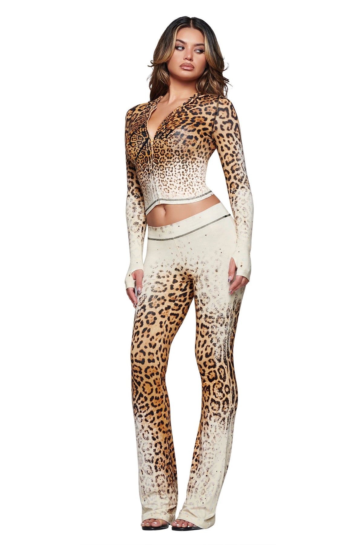 A woman stands confidently against a white background, wearing the I.AM.GIA BLARE TRACKPANT in Cream Faded Panthera—a fitted, long-sleeve leopard-print crop top paired with matching cheetah print track pants. Her hair falls in loose waves and she completes the look with open-toe heels.