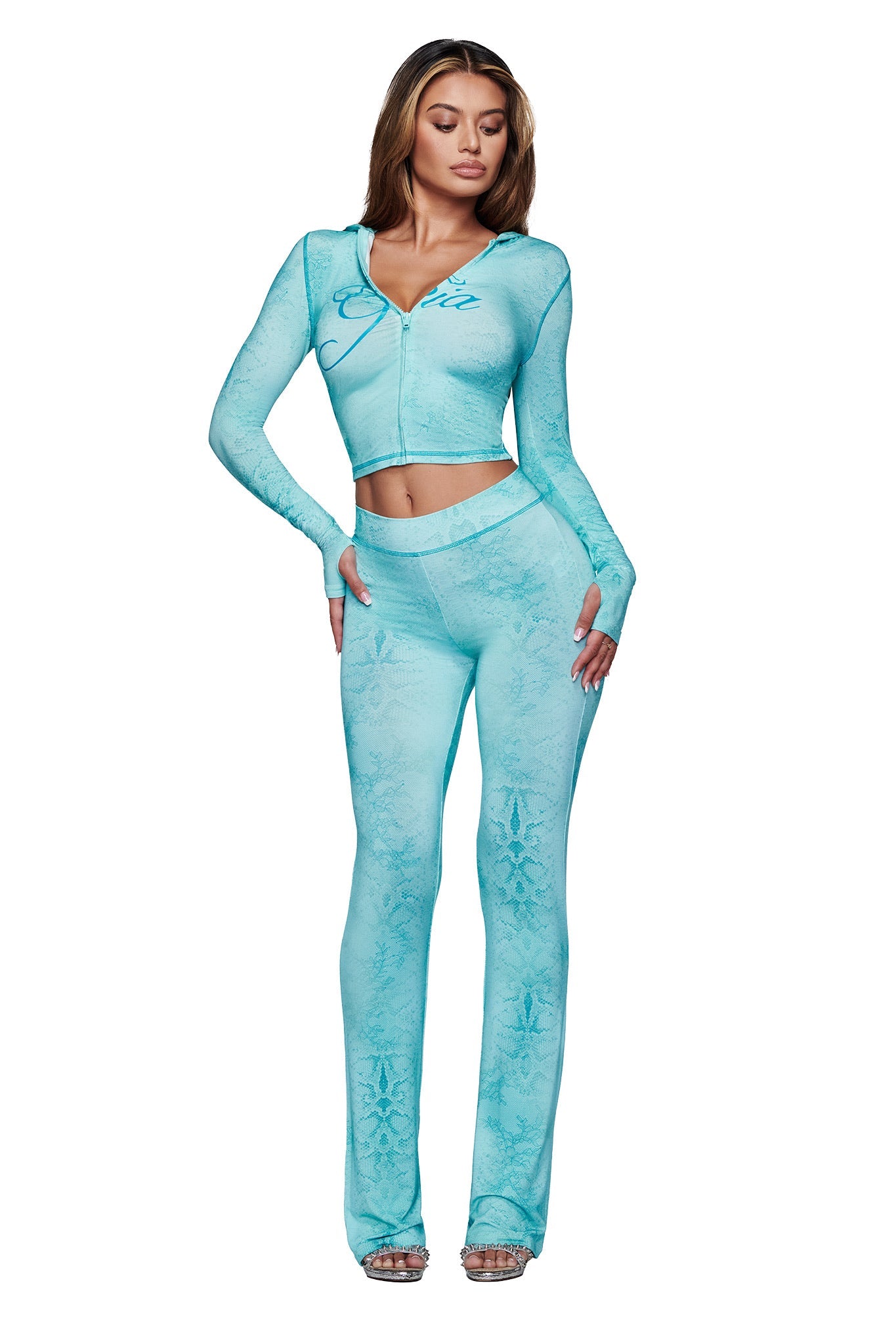 A woman wears I.AM.GIA’s light blue, fitted, long-sleeve zip-up top with the BLARE TRACKPANT - TEAL SERPENTINE featuring a flared leg. She pairs the outfit with silver high-heeled sandals and poses against a white background.