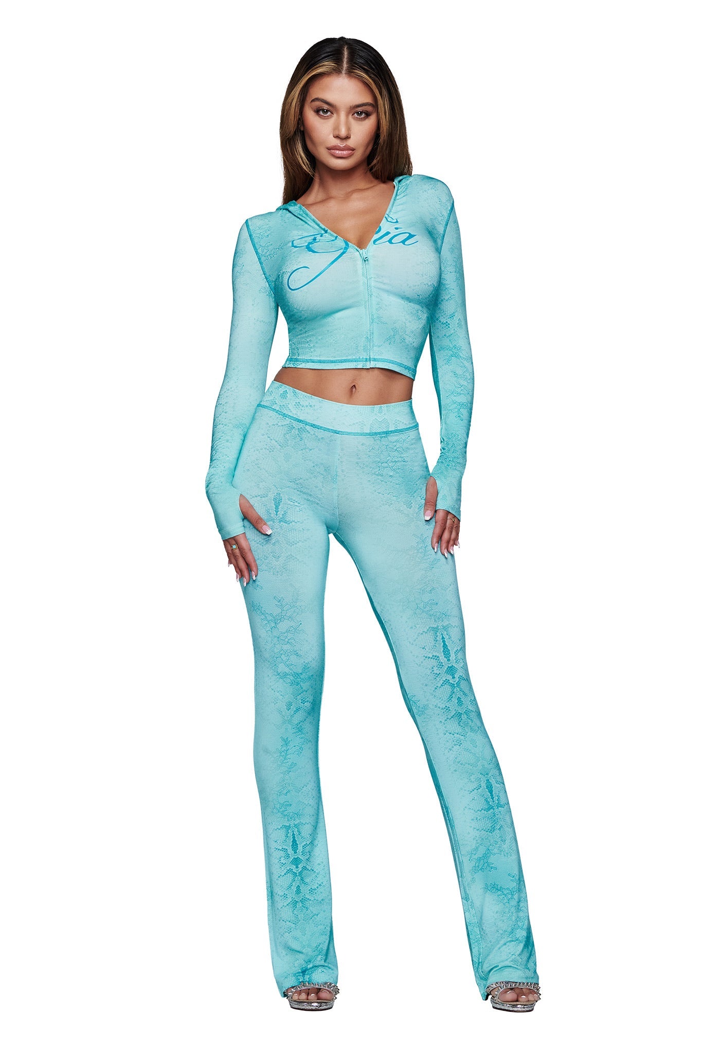 A woman faces forward wearing the I.AM.GIA BLARE TRACKPANT - TEAL SERPENTINE, styled with a matching zip-up crop top. She has long straight hair and silver high-heeled sandals. The trackpants feature a flared leg and subtle pattern.