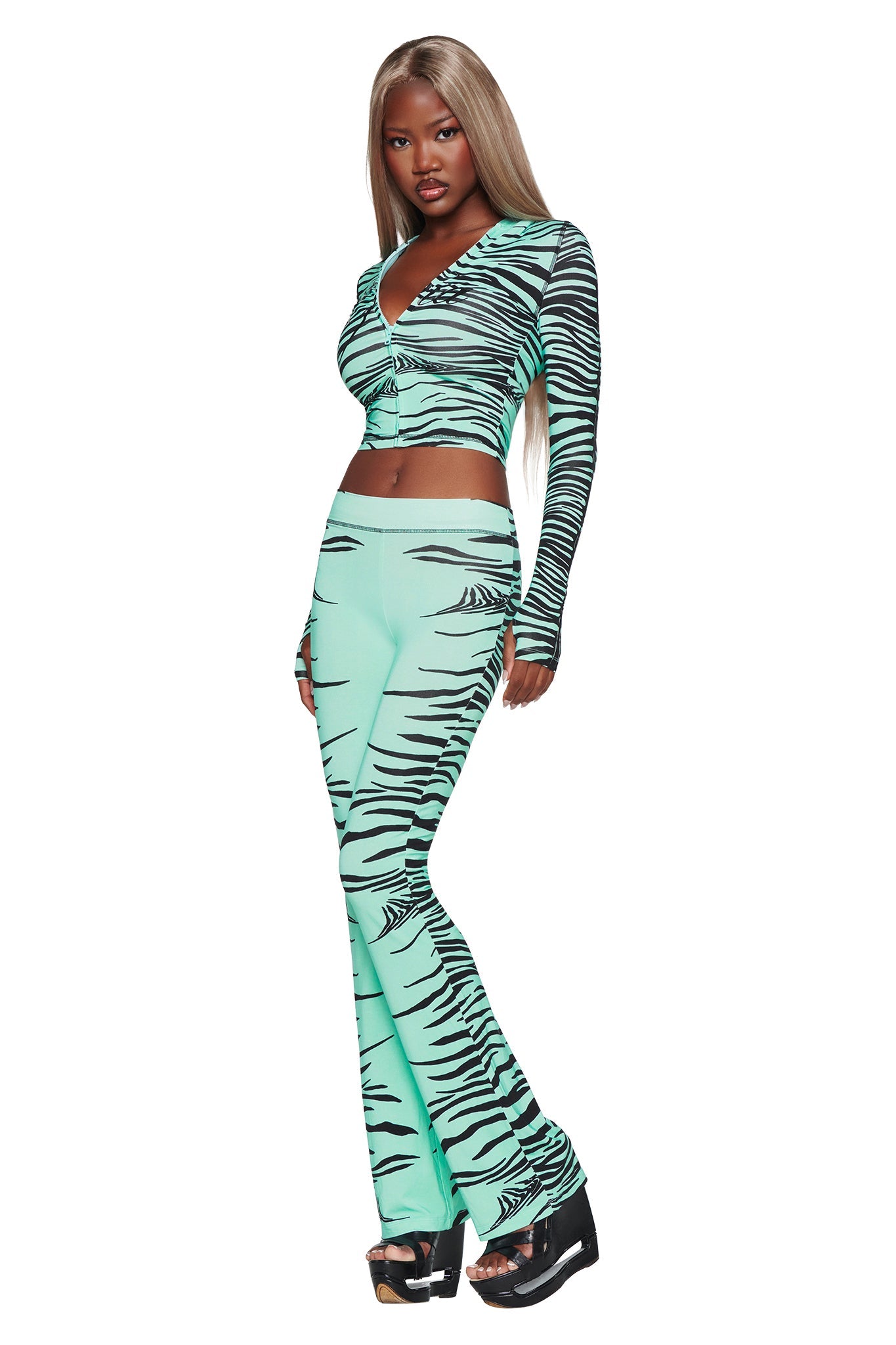 A woman poses against a white background wearing the I.AM.GIA BLARE TRACKPANT in Aqua Fine Line Zebra, styled as a matching set with a long-sleeve crop top and flared leg trackpants, completed with black platform heels.