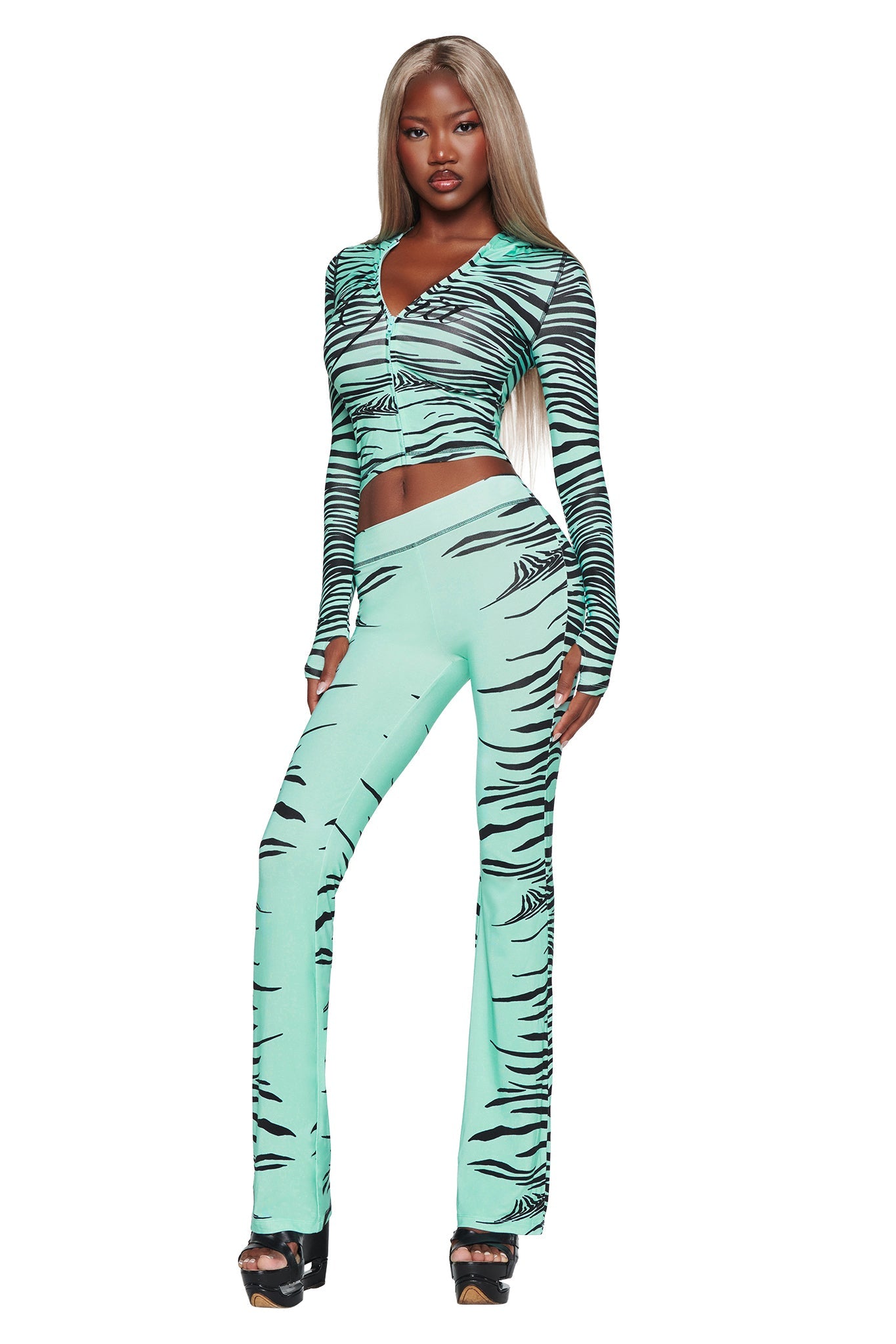 A woman with long blonde hair stands confidently against a white background, wearing the I.AM.GIA BLARE TRACKPANT in Aqua Fine Line Zebra print paired with a matching crop top and black platform heels.