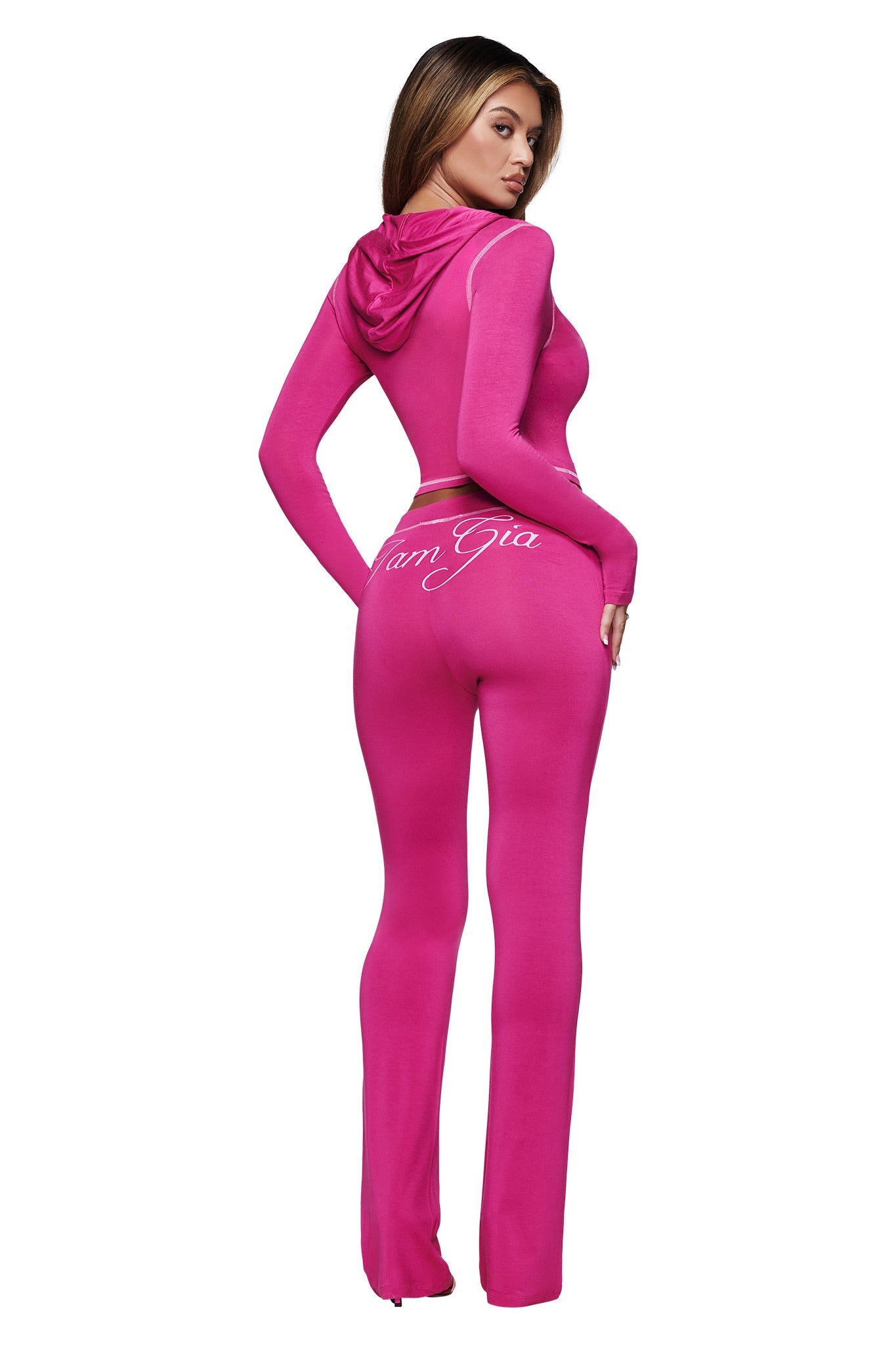A woman poses with her back to the camera, wearing I.AM.GIA’s BLARE TRACKPANT in raspberry. The fitted pants have flared legs and white script text reading "IamGia" across the back.
