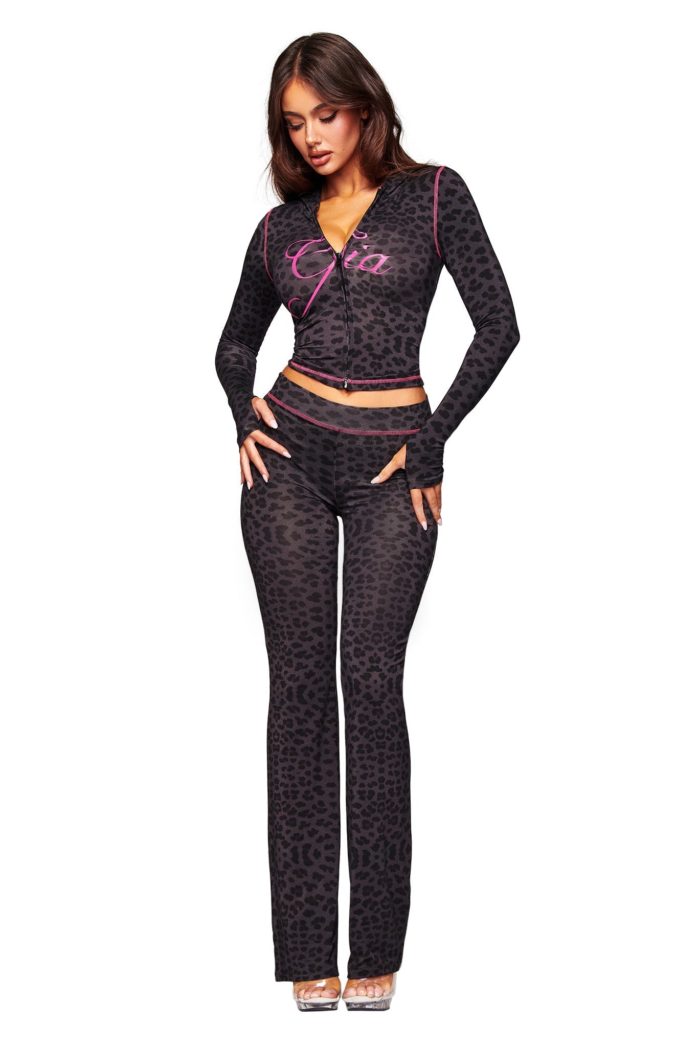 A woman wears I.AM.GIA’s fitted BLARE TRACKPANT - PANTHER set, featuring a long-sleeve zip-up top and flared panther-print pants. She has long brown hair, clear heels, and poses looking down with hands on hips.