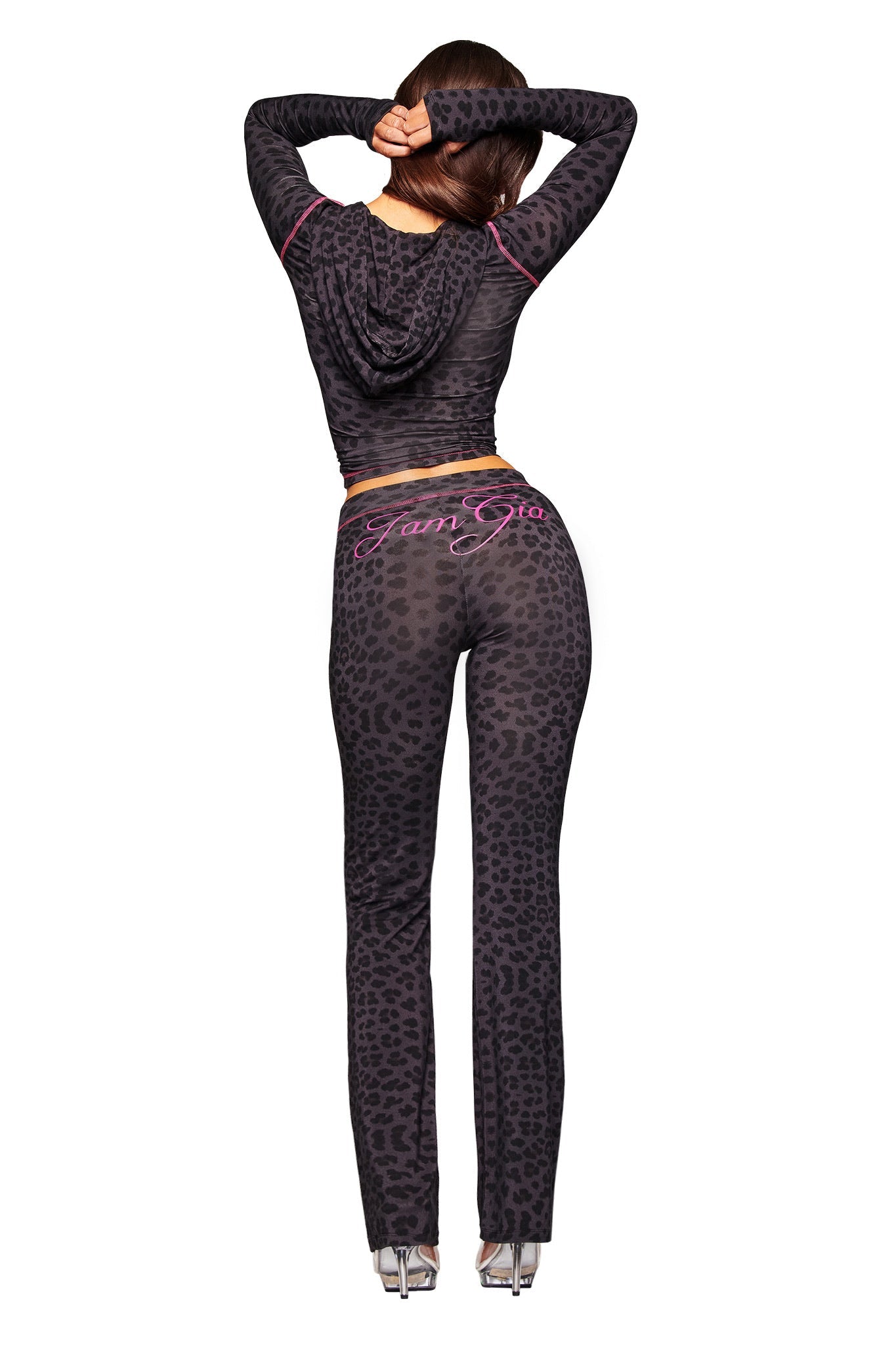 A woman faces away from the camera with arms raised, wearing I.AM.GIA’s fitted black and gray BLARE TRACKPANT - PANTHER and clear heels. "FAMGIA" in pink appears on the back of her pants.