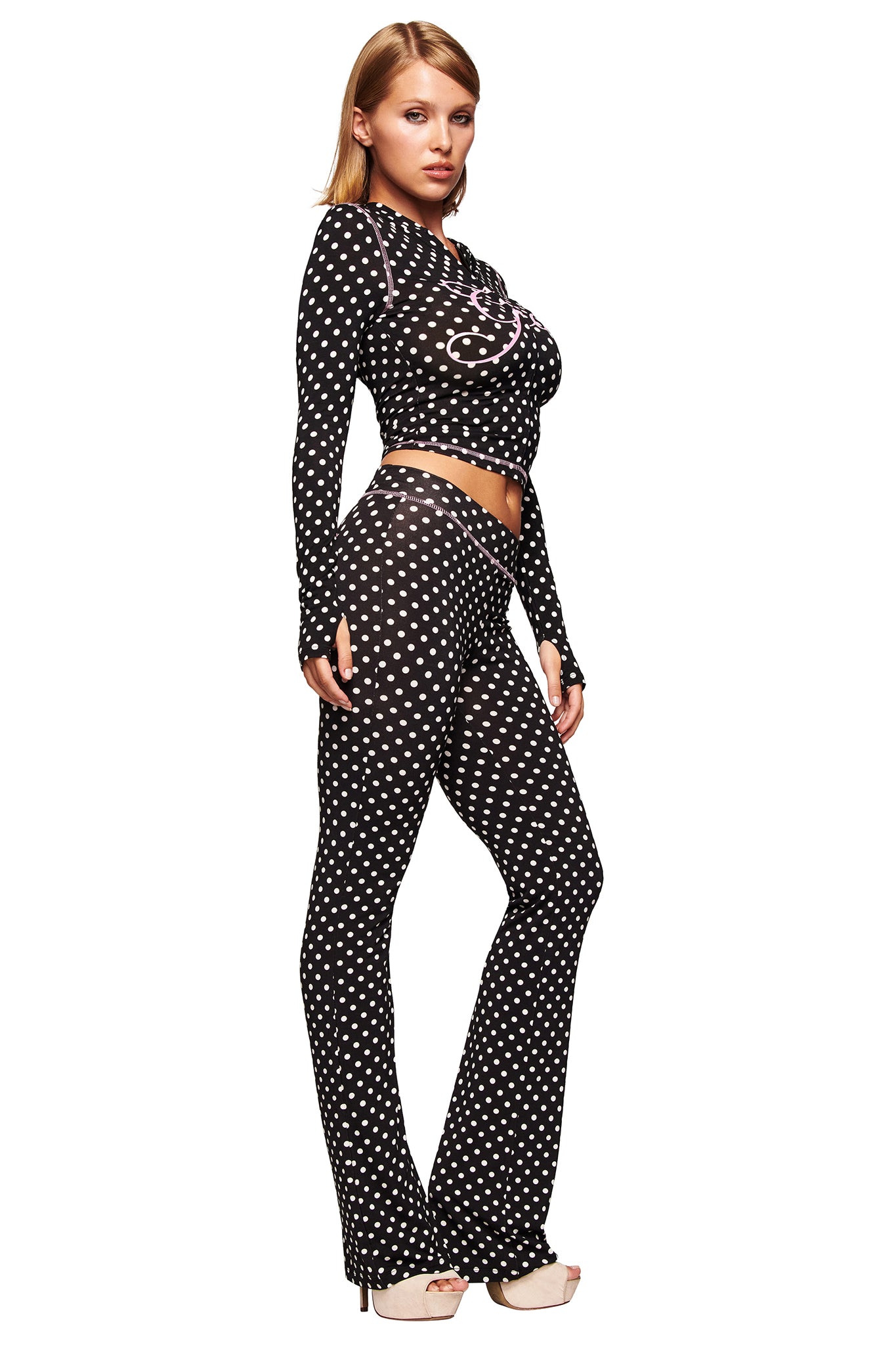 A woman poses in an I.AM.GIA BLARE TRACKPANT - BLACK POLKA DOT and matching sheer long-sleeve top, both with white polka dots. She wears nude heels and has straight, shoulder-length blonde hair against a white background.