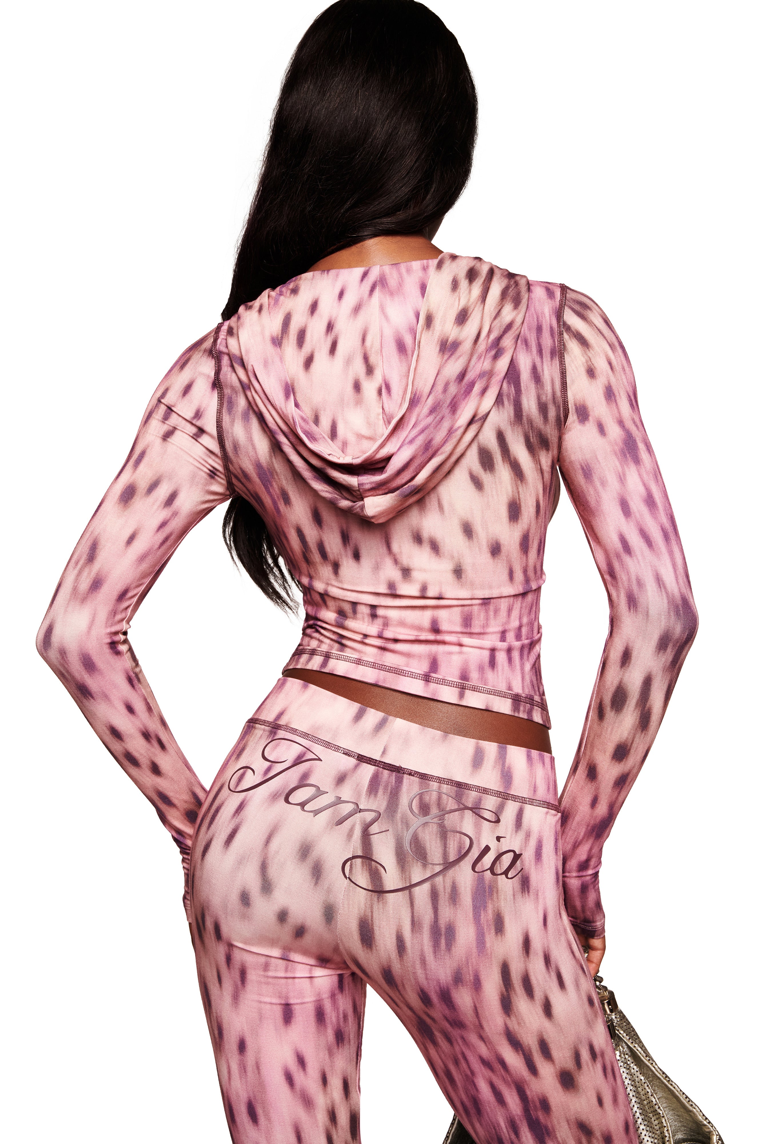A woman with long dark hair is seen from behind, wearing a fitted I.AM.GIA BLARE HOODIE in Pink Blurred Cheetah and matching leggings with "IamGia" on the back, holding a silver handbag in her left hand.