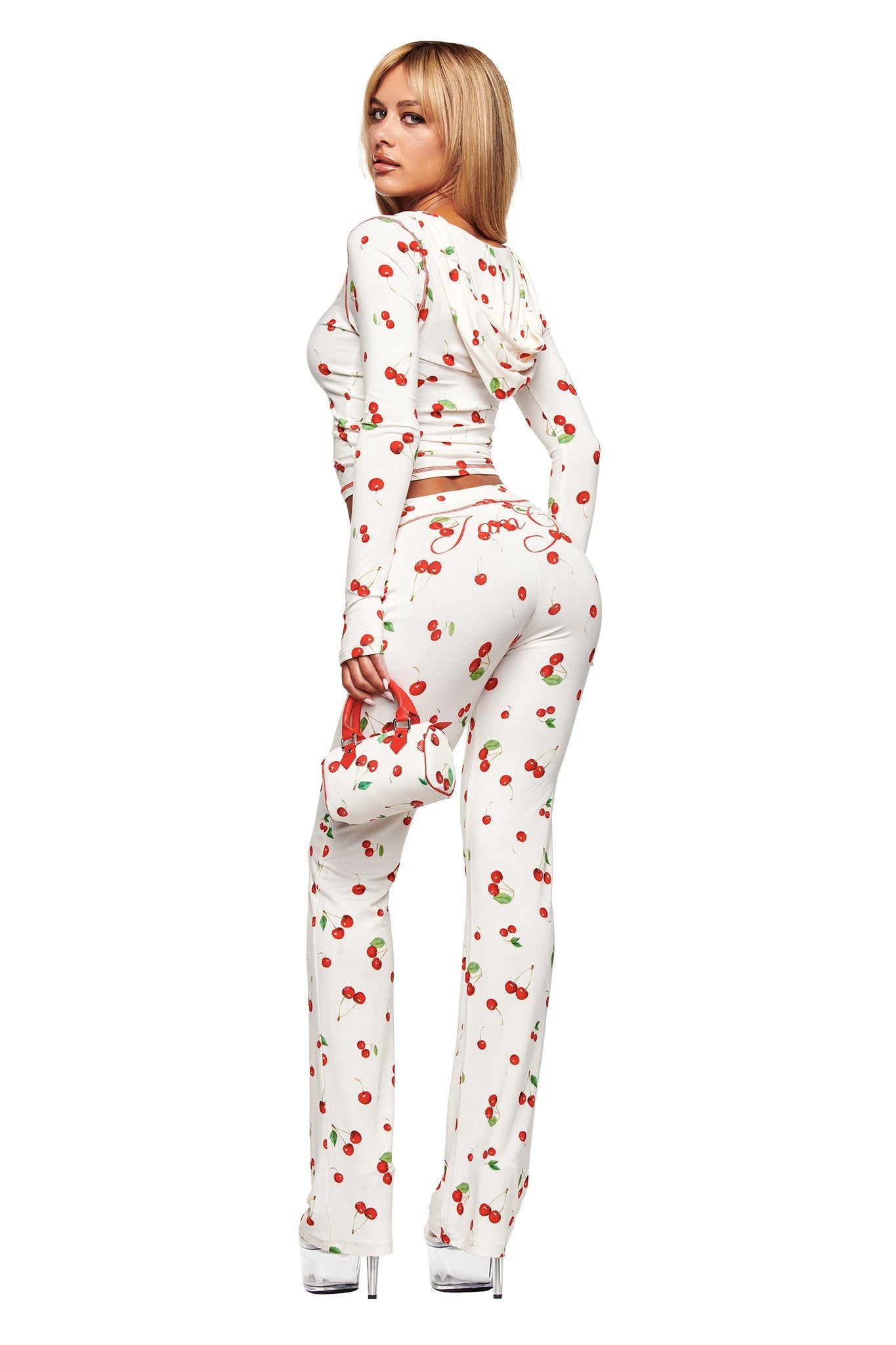 A woman faces away, turning her head to the camera. She wears a white hooded crop top and I.AM.GIA’s BLARE TRACKPANT in Cherry on Top, paired with clear high heels and a matching small purse.