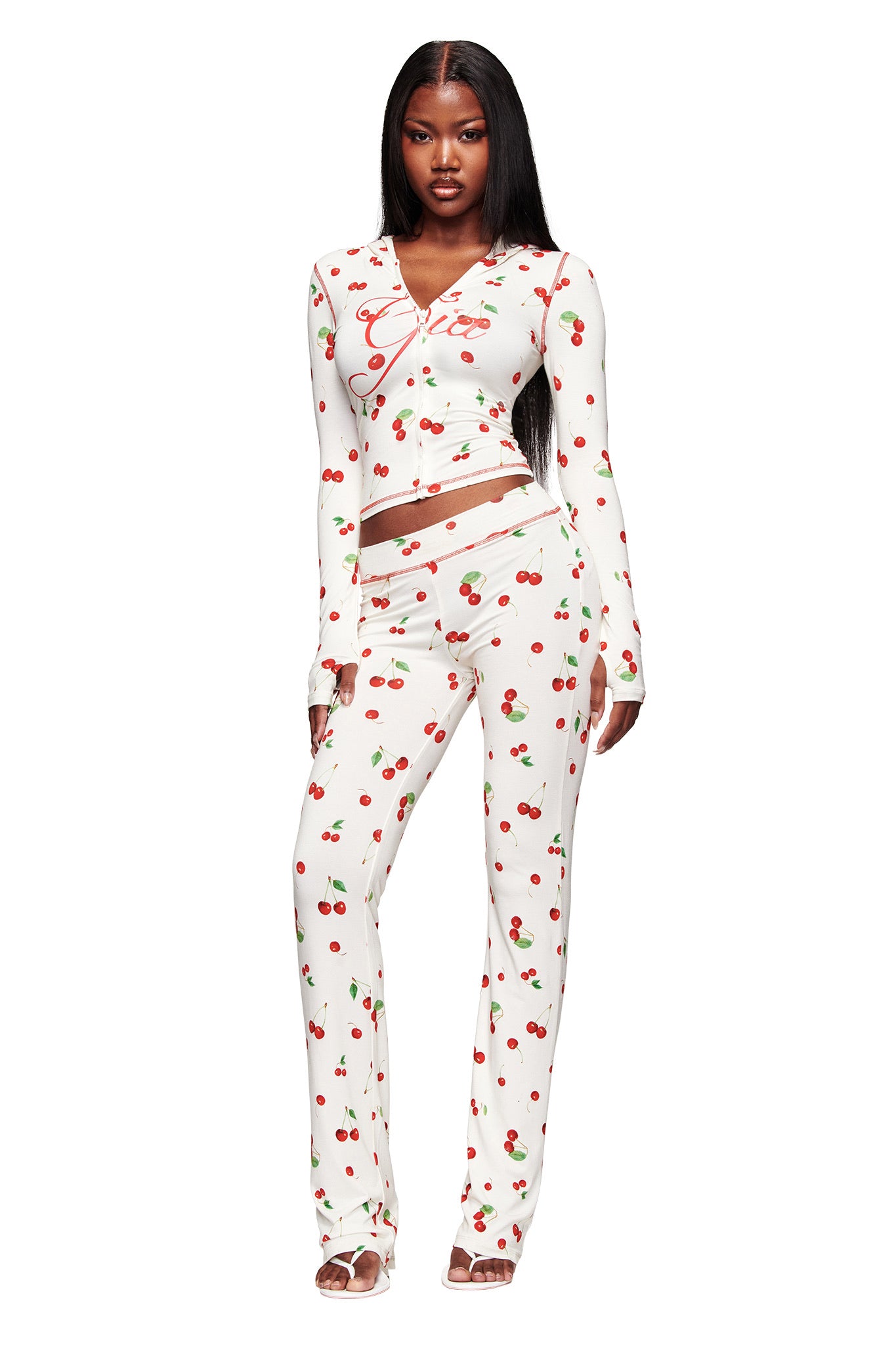 A woman wears the I.AM.GIA BLARE TRACKPANT in Cherry on Top, made from soft bamboo. She pairs it with a white long-sleeve top and open-toe heels, her long straight hair down, standing against a white background with a neutral expression.