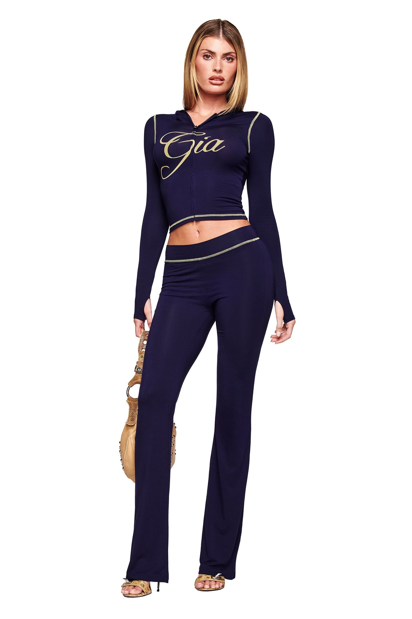 A woman stands confidently in I.AM.GIA’s BLARE TRACKPANT - NAVY, a two-piece set with long sleeves and flared pants featuring “Gia” in gold on the top. She accessorizes with a tan handbag and gold high-heeled sandals.