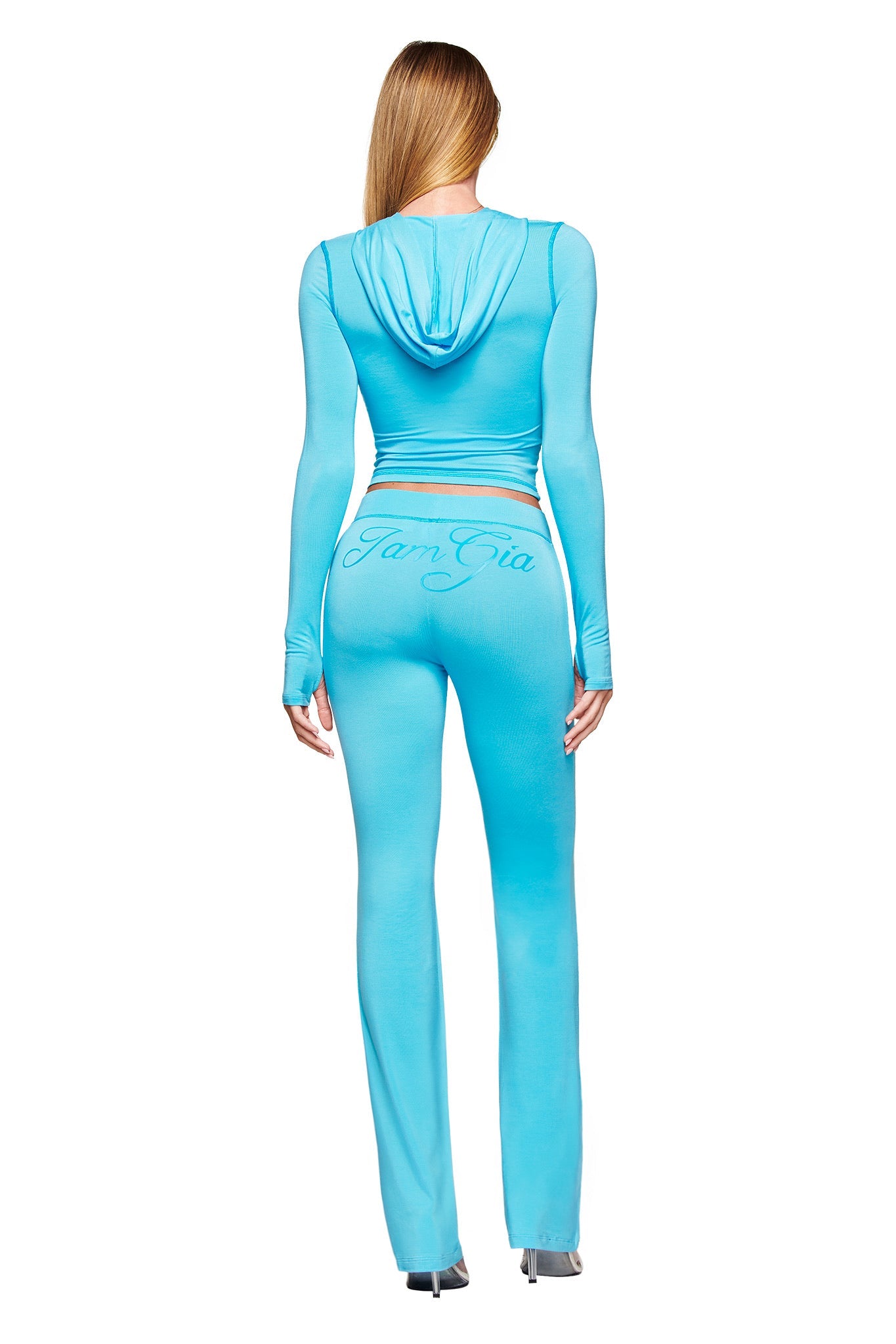 A woman with long straight hair is seen from behind against a white background, wearing a fitted light blue hooded top and I.AM.GIA's BLARE TRACKPANT in neon blue, featuring the brand logo on the back.