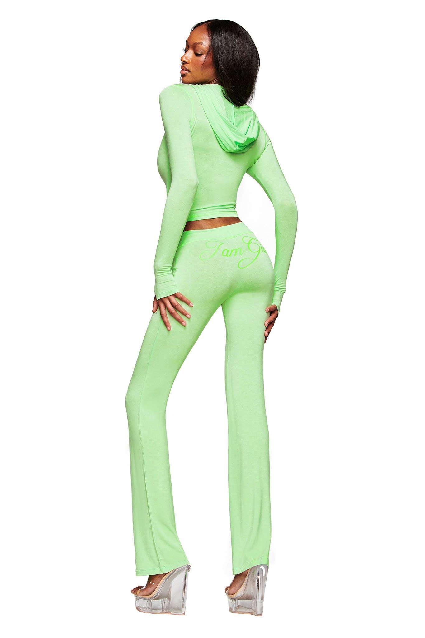 A person poses against a white background in I.AM.GIA's BLARE TRACKPANT in neon green with "I am G" on the back, styled with a fitted light green hooded crop top and platform heels.