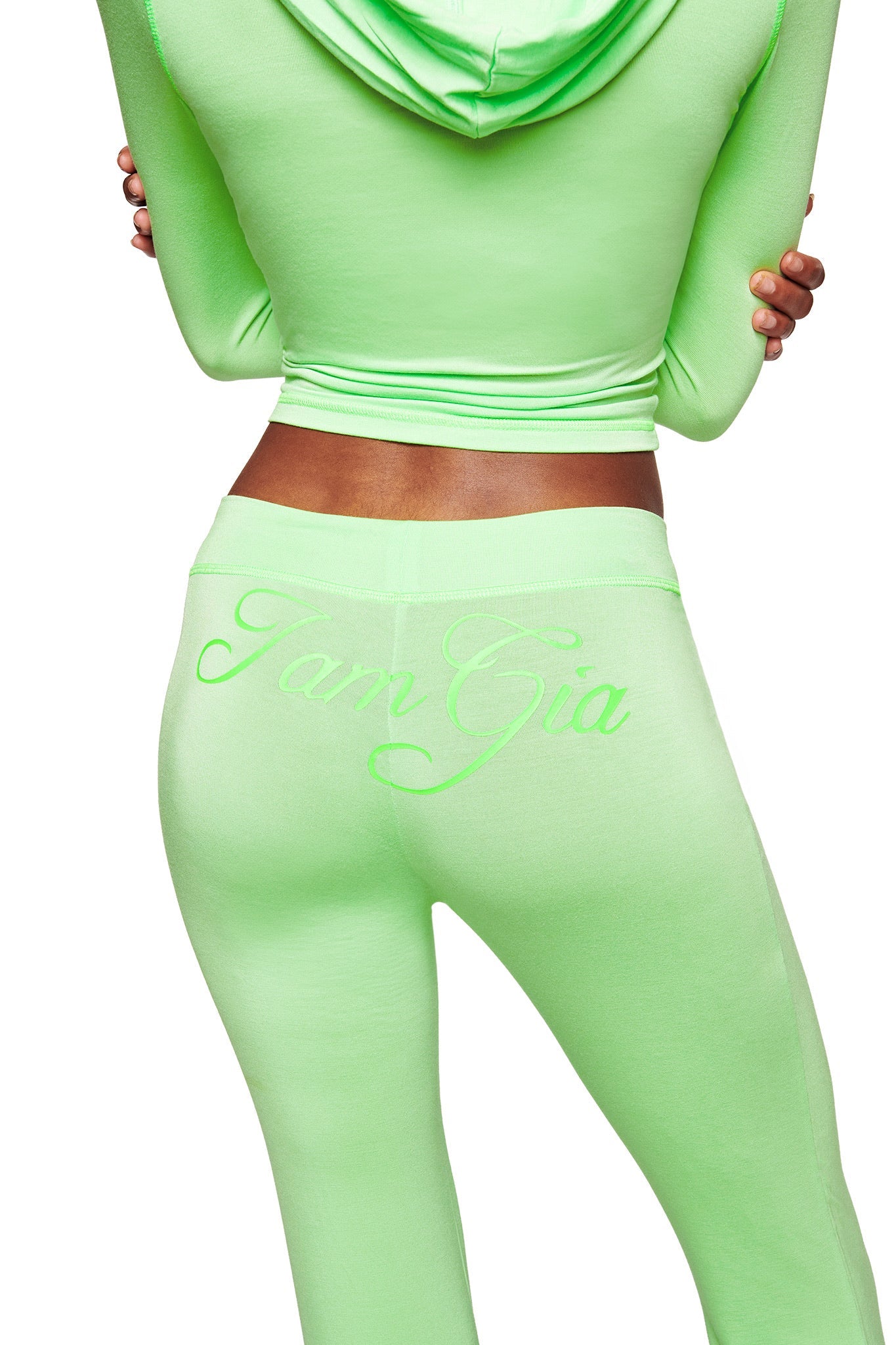 A person is seen from the back wearing a bright green fitted hoodie and I.AM.GIA's BLARE TRACKPANT in neon green, featuring cursive "I.AM.GIA" embroidery on the flared trackpants for a bold, standout look.