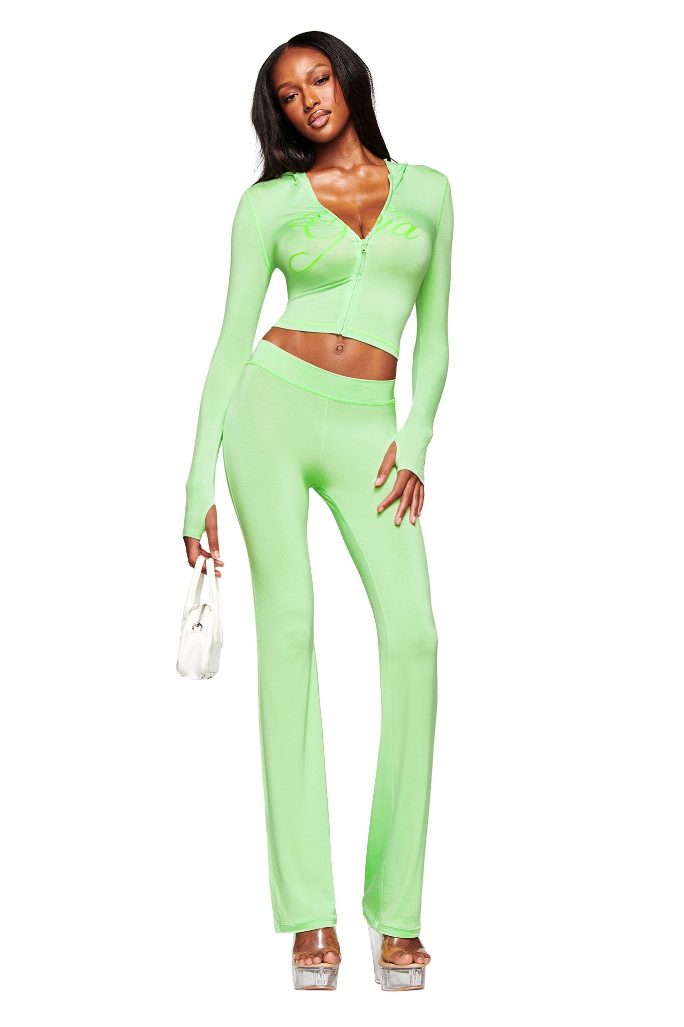 A woman models the I.AM.GIA BLARE TRACKPANT in neon green with a matching zip-up crop top, paired with a small white handbag and high platform heels, posed against a plain white background.
