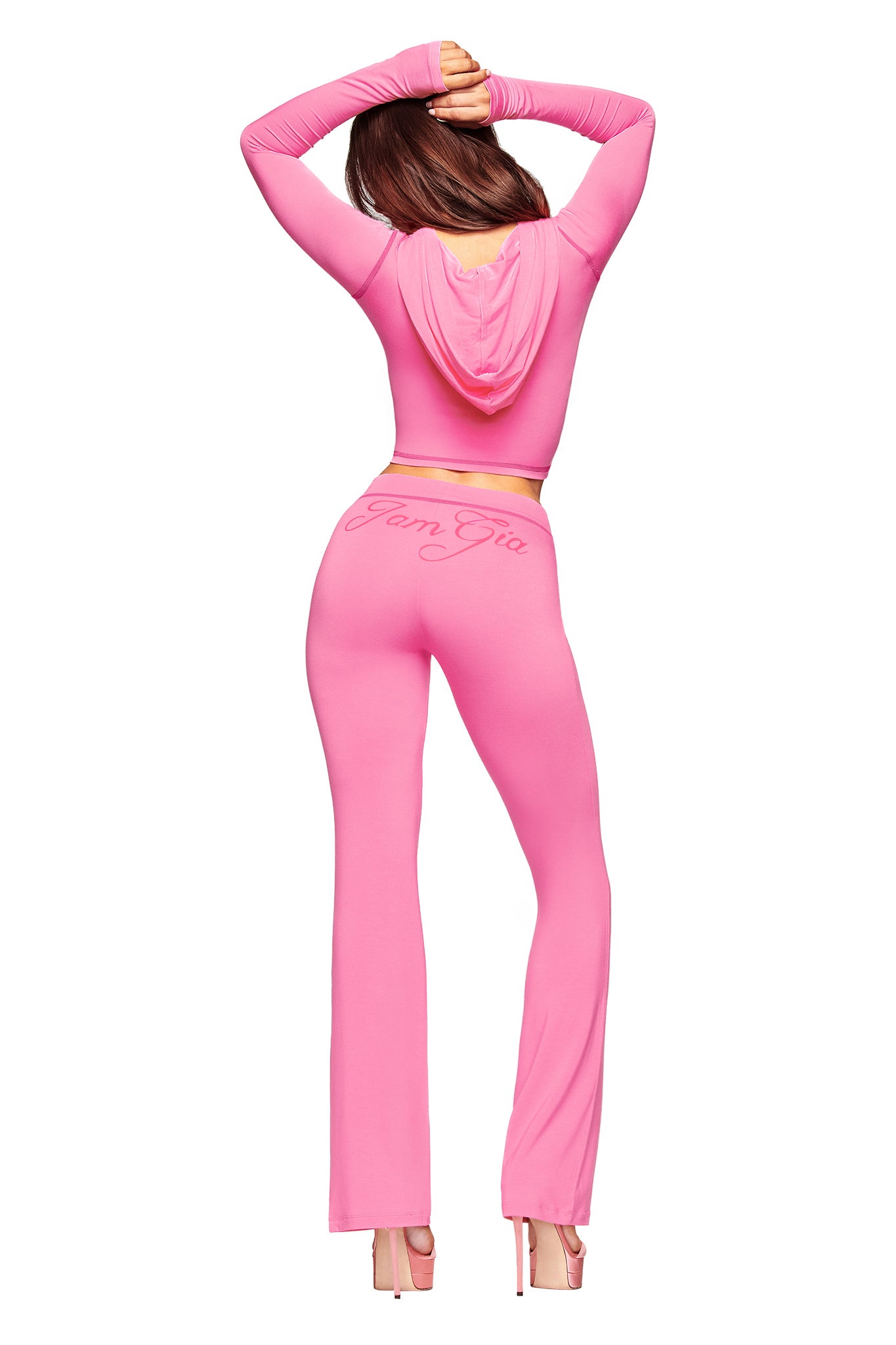 A person in the I.AM.GIA BLARE TRACKPANT - NEON PINK set and matching hoodie with “Pam Gia” on the back stands on high platform heels, arms raised above their head, facing away.