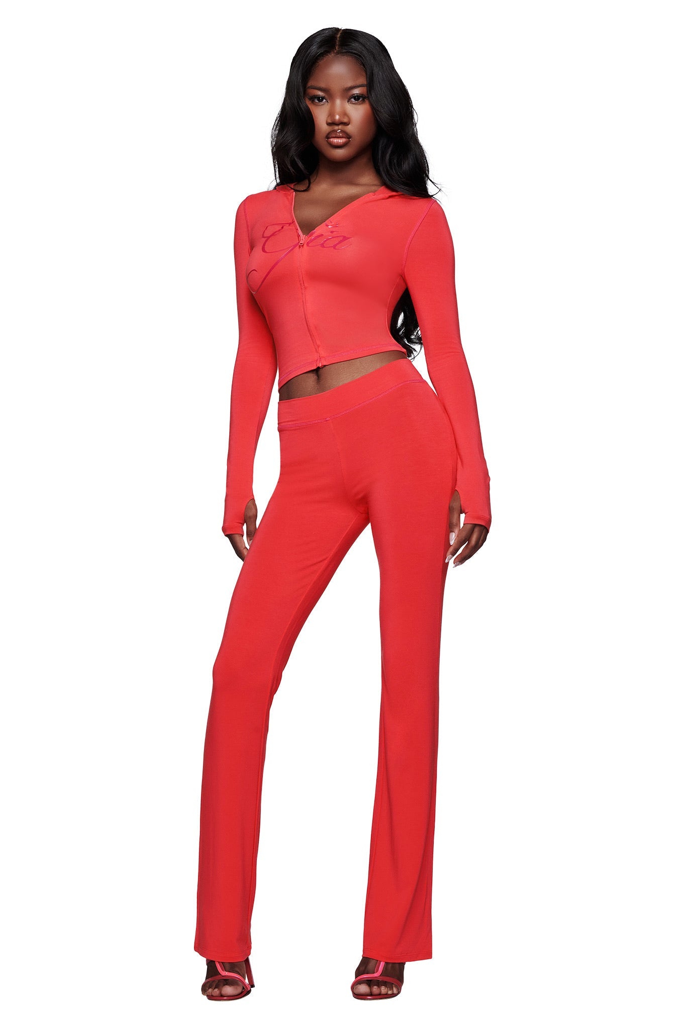 A woman stands confidently in the I.AM.GIA BLARE TRACKPANT - PAPAYA, a fitted, bright red two-piece with flared legs and a mid-rise waist, paired with matching red heels against a plain white background.