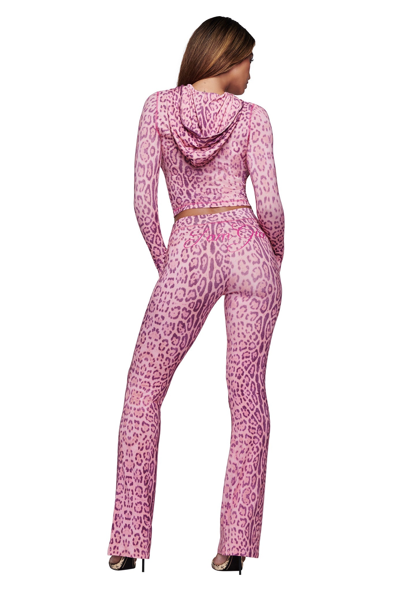 A person with long hair stands facing away, wearing the I.AM.GIA BLARE TRACKPANT - PINK PANTHERA and a matching hooded crop top. The outfit features a snug fit, flared legs, and is styled with high heels.