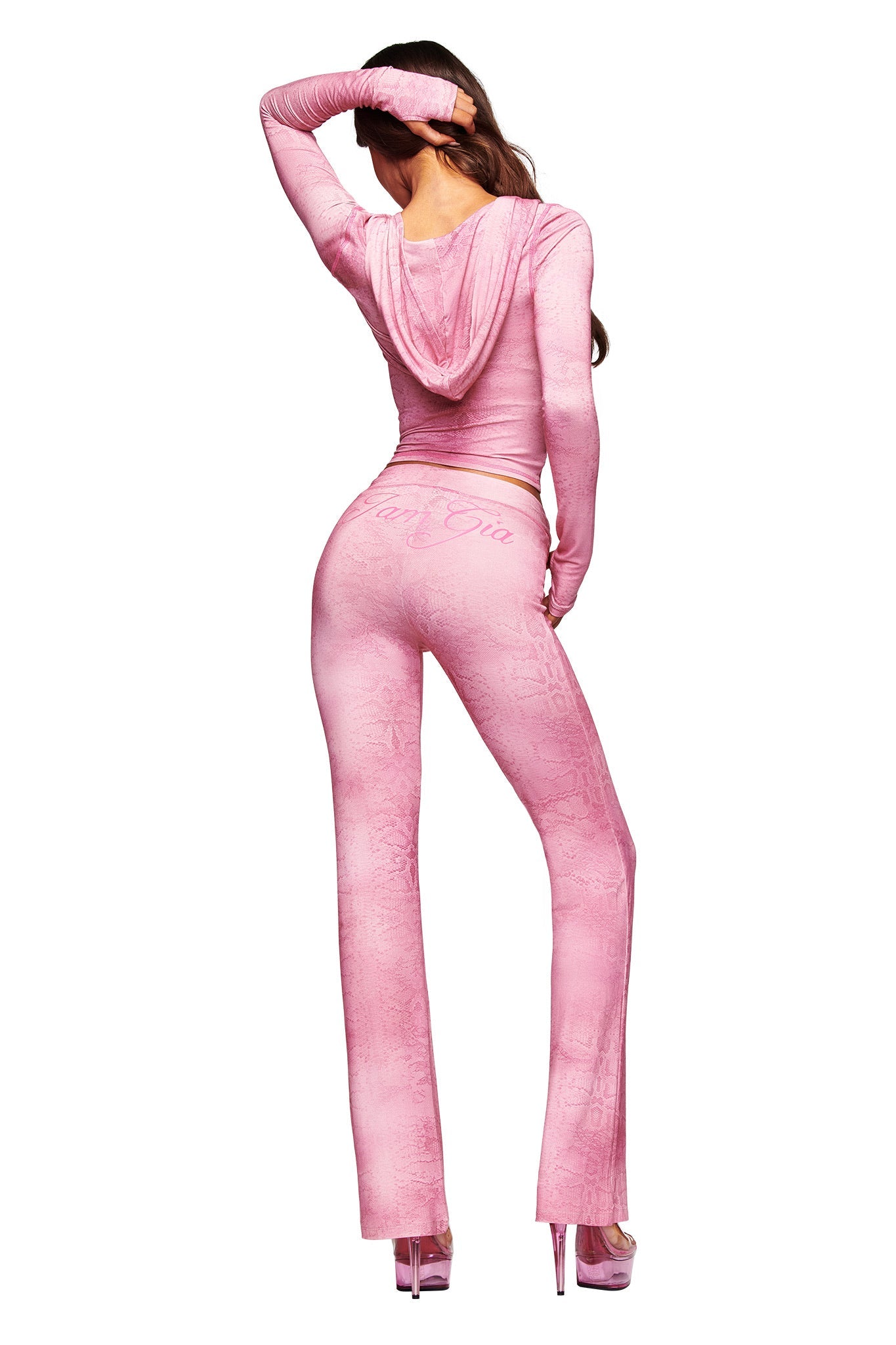 A woman faces away, wearing the I.AM.GIA BLARE TRACKPANT in Light Pink Serpentine print with a flared leg, paired with a matching fitted hooded top and platform heels. One arm is raised, her long hair flowing down her back.