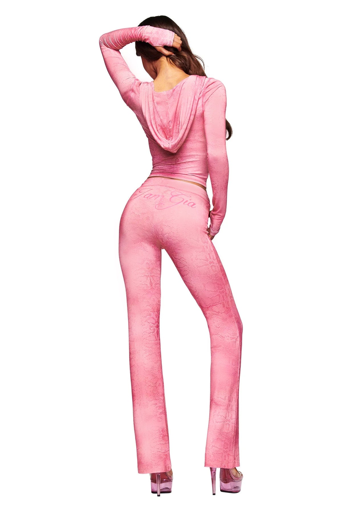 A woman stands with her back to the camera, wearing the I.AM.GIA BLARE TRACKPANT in Light Pink Serpentine print, paired with a matching fitted hooded top and high heels. Her left arm is raised to rest on her head, and her long hair cascades down her back.