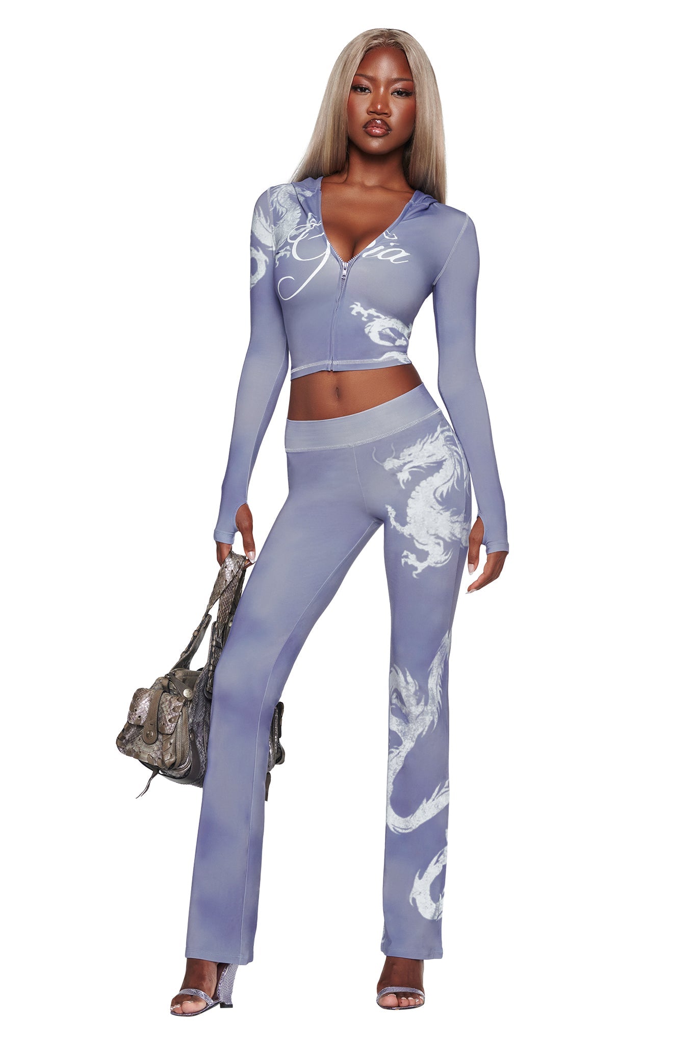 A woman stands confidently against a white background, wearing the I.AM.GIA BLARE TRACKPANT in Steel Blue Dragon paired with a fitted, light purple two-piece outfit featuring white swirling patterns. She completes the look with heels and a handbag.