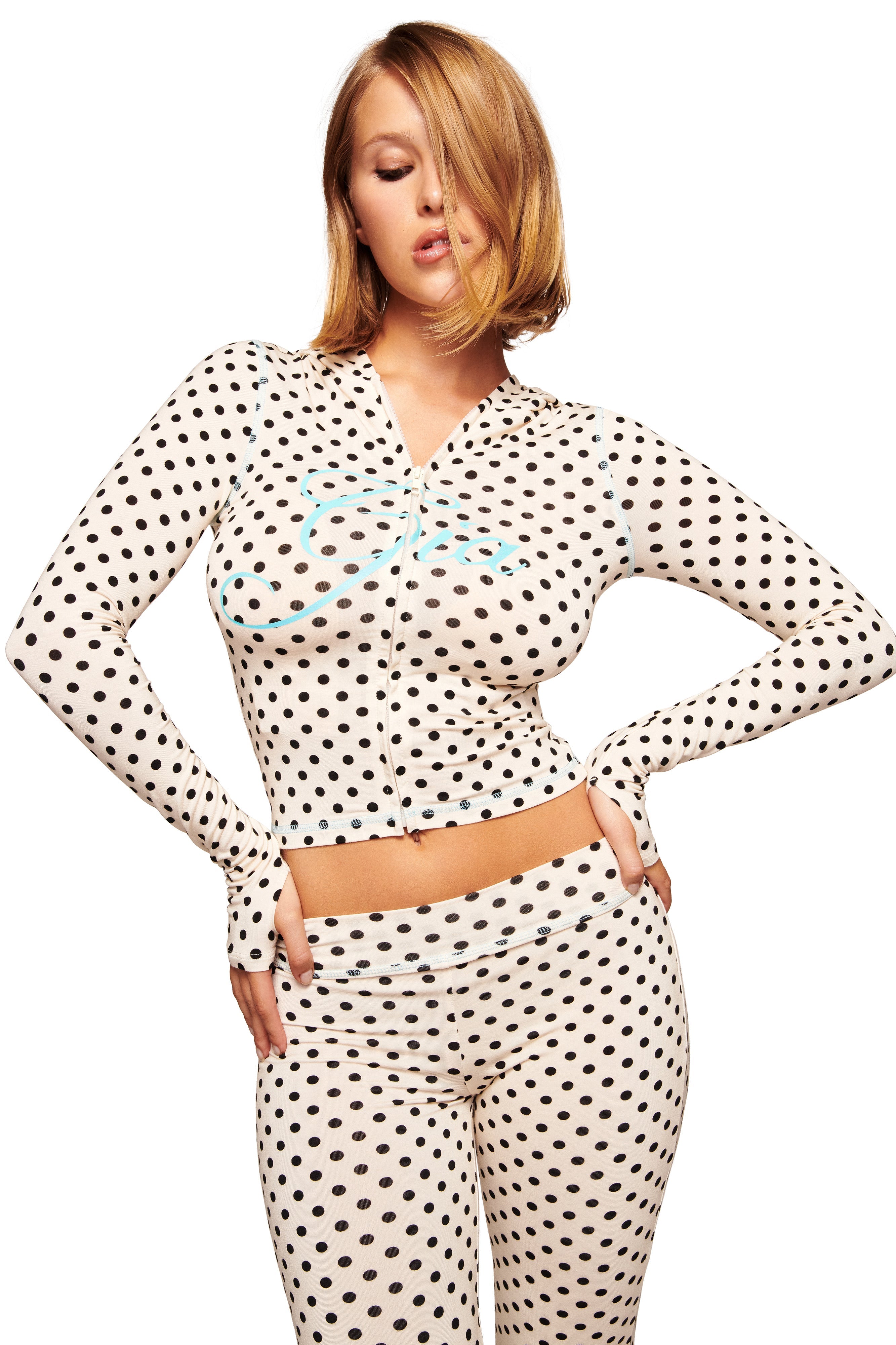 A blonde woman models the I.AM.GIA BLARE HOODIE in cream polka dot, striking a pose with one hand on her hip and her head tilted, some hair falling over one eye, against a white background.
