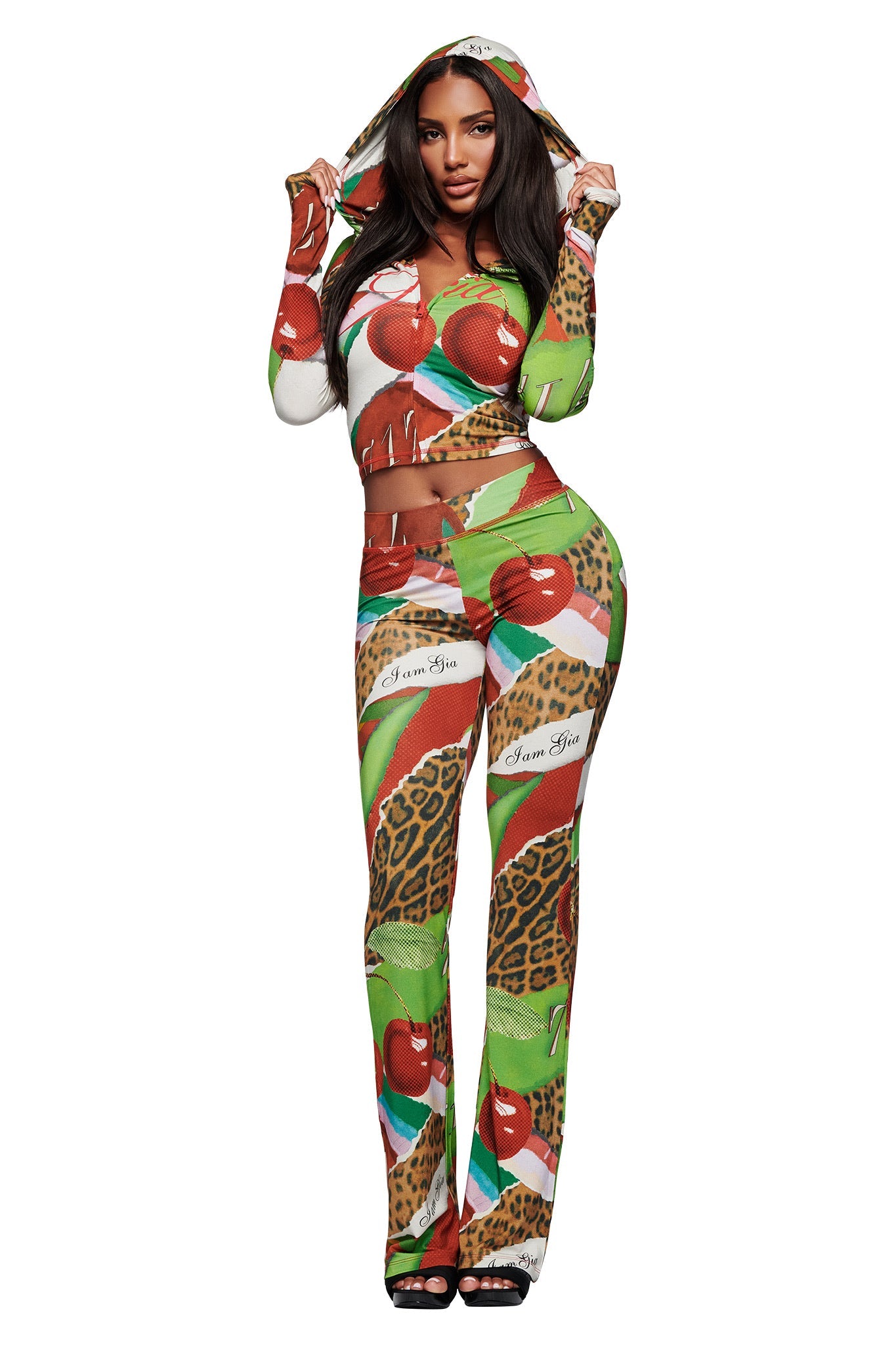 A woman wears the I.AM.GIA BLARE HOODIE in Wild Cherry, a colorful form-fitting two-piece with a hooded top and matching pants featuring leopard spots, cherries, and green leaves, standing against a white background.