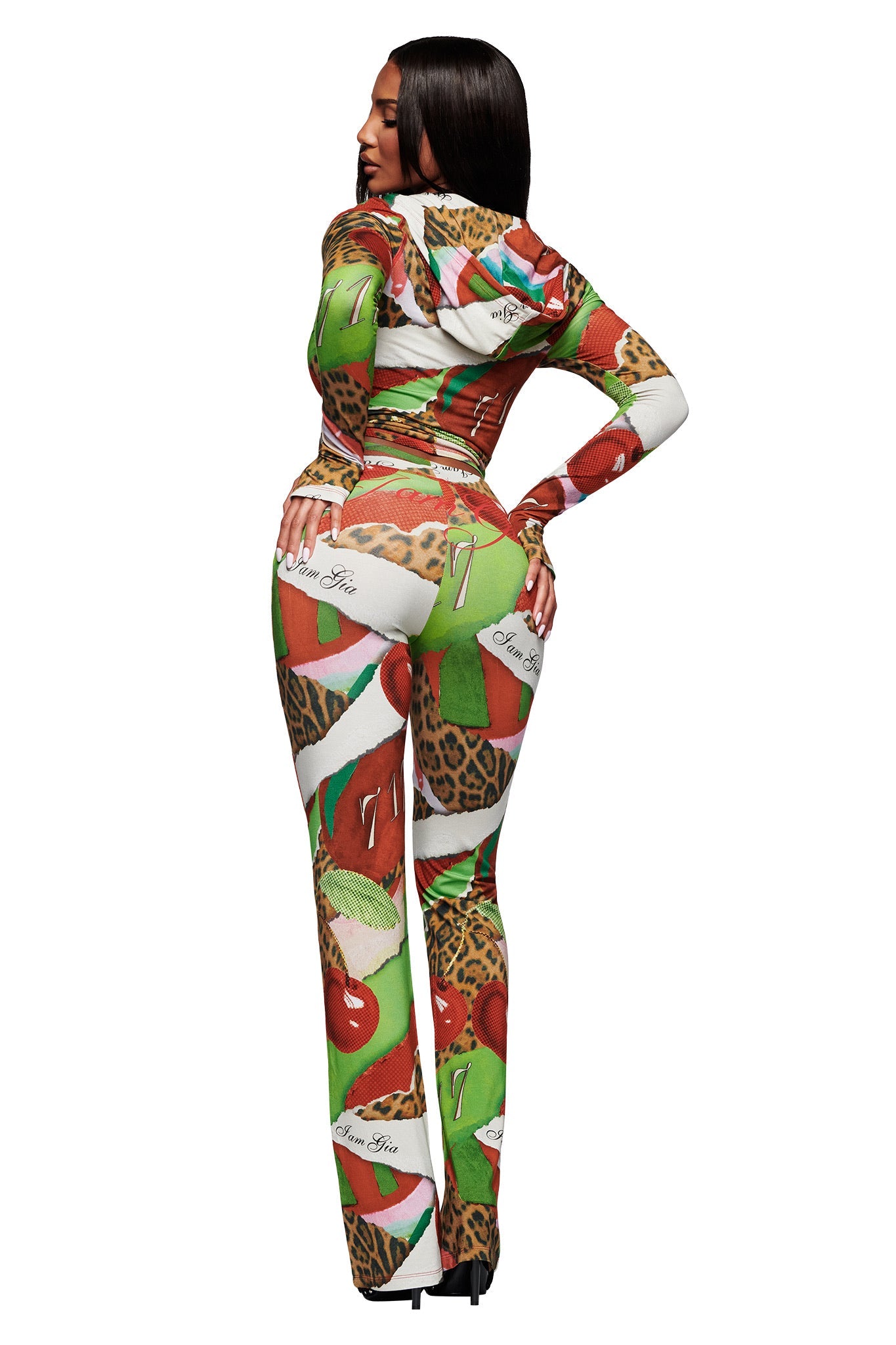 A woman poses with her back to the camera, wearing I.AM.GIA's BLARE TRACKPANT in Wild Cherry, featuring bold leopard patterns, green and red shapes, and white sections with cursive writing.