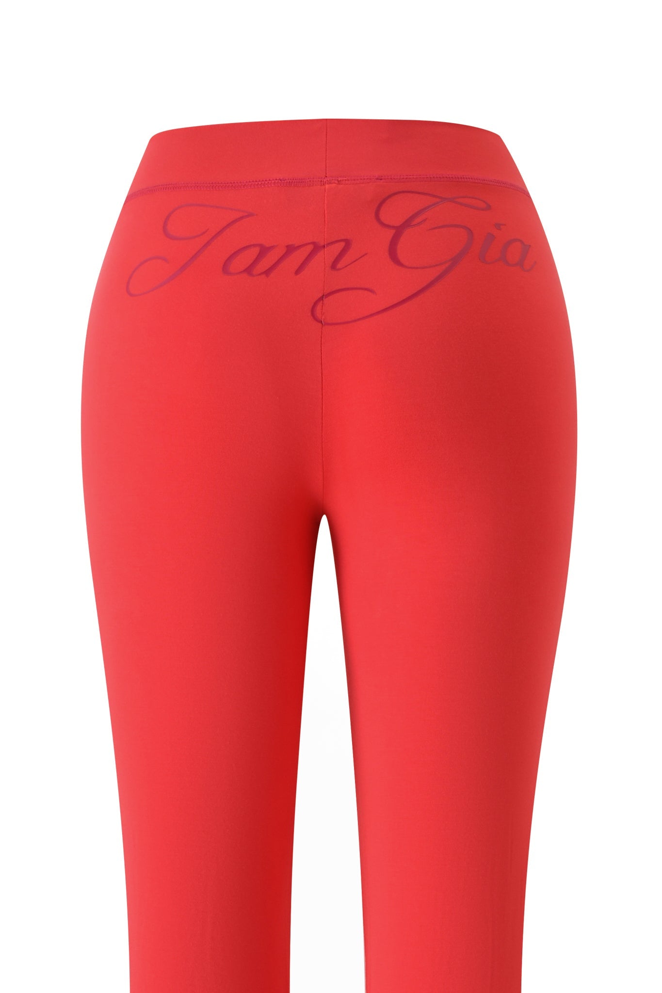 The BLARE TRACKPANT - PAPAYA by I.AM.GIA is shown from the back, with “I.AM.GIA” in cursive on the waistband. These bright red pants have a mid rise waist and flared legs, set against a plain white background.