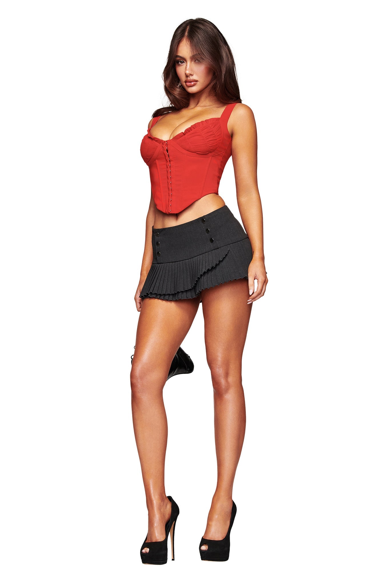 A woman poses in the I.AM.GIA BRI CORSET in rich red, styled with a black pleated mini skirt and black high heels, against a plain white background.