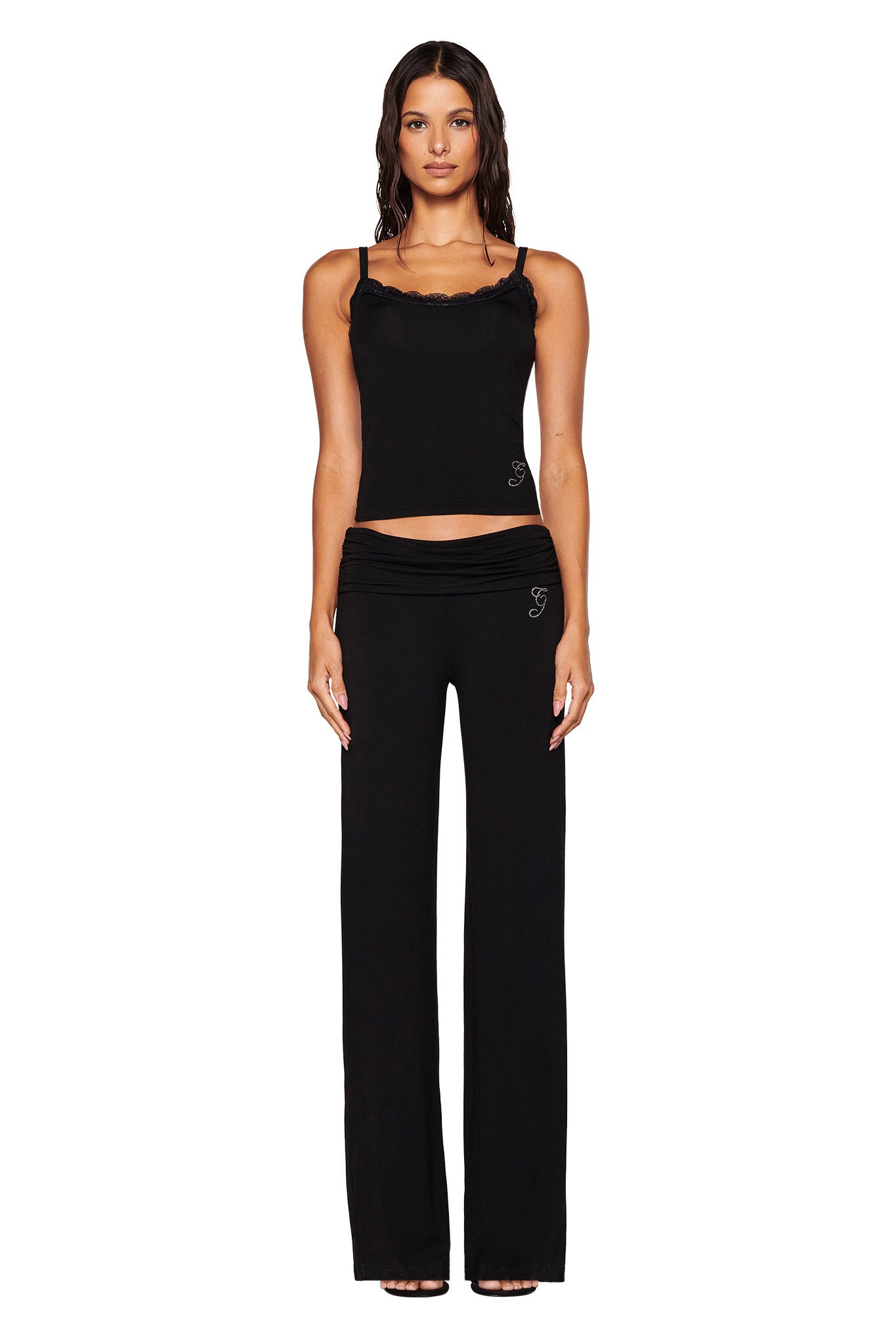 A woman stands facing forward in the stylish ERIN TOP - BLACK by I.AM.GIA, featuring an elegant lace trim, paired with black wide-leg pants. Her long, wavy dark hair cascades against the plain white background, showcasing the effortless grace of her look.