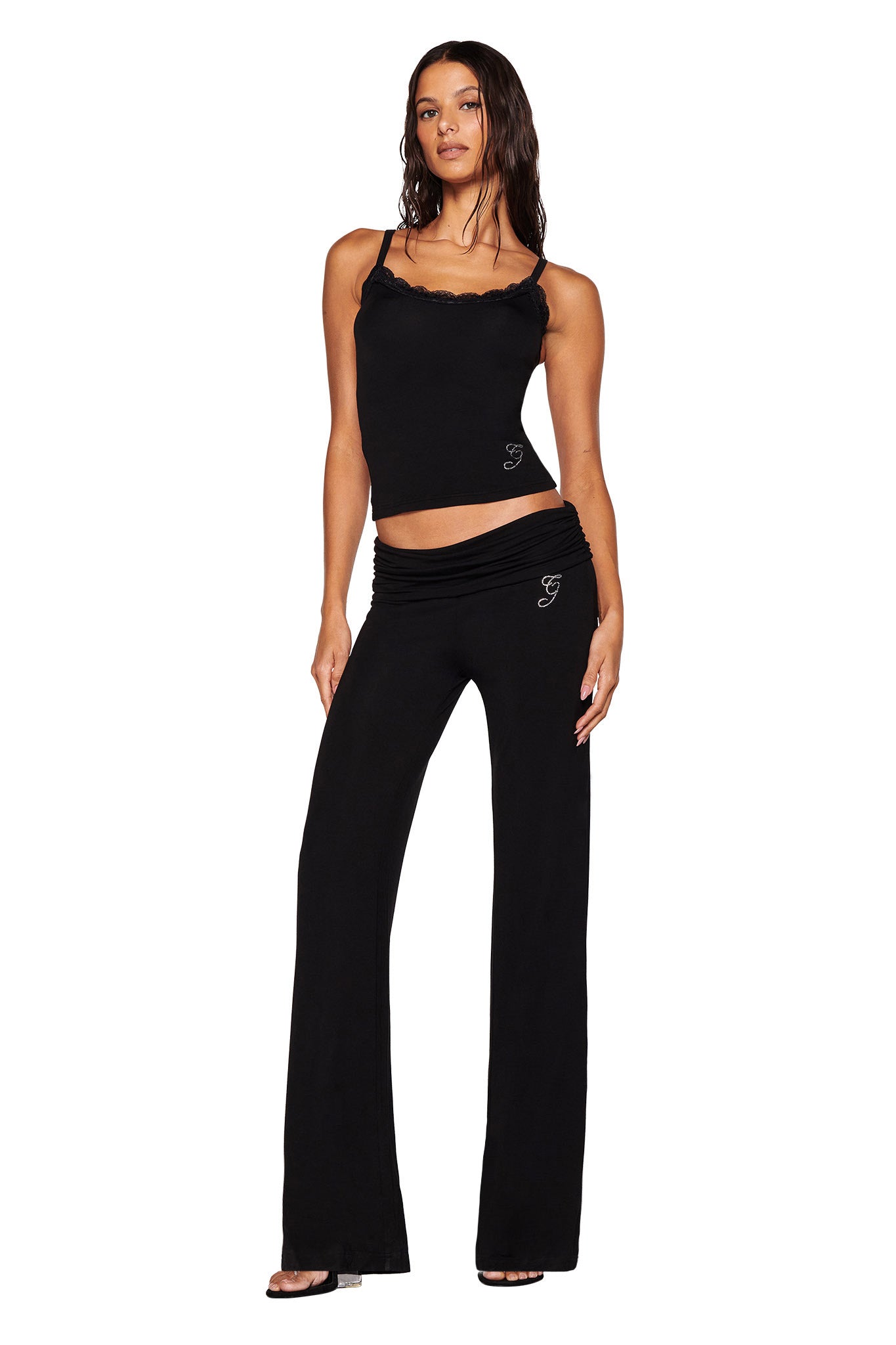 Wearing a chic black spaghetti strap top with ruffled edges, the individual stands confidently in the matching I.AM.GIA ERIN PANT - BLACK. Their sleek wet-look hair enhances the open-toed black sandals, while delicate diamante accents provide a hint of sparkle against the plain white backdrop.