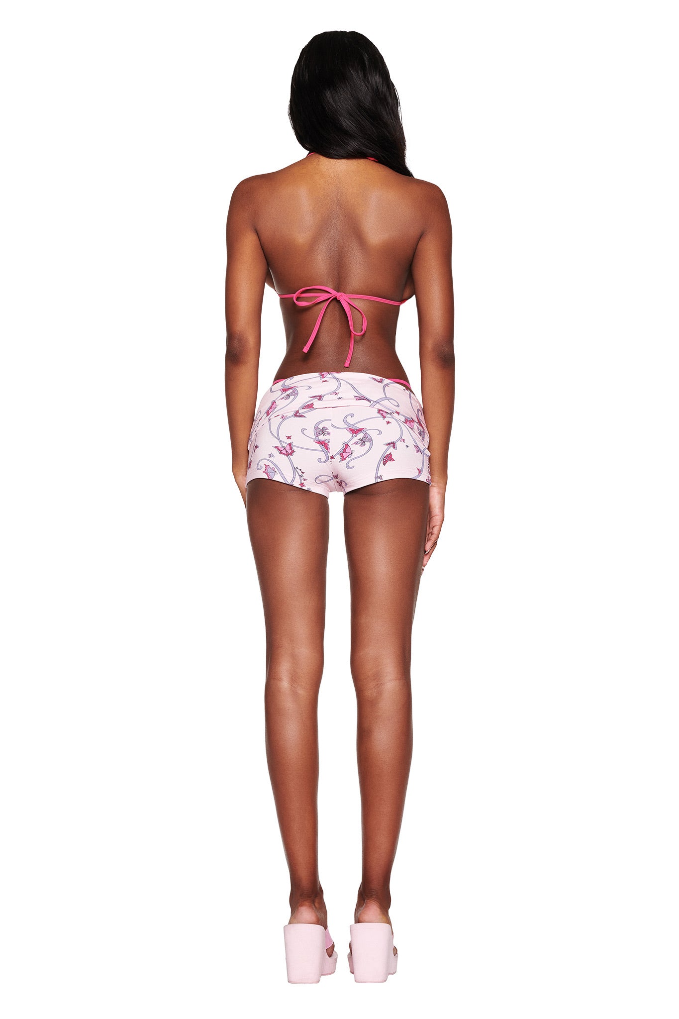 A person with long hair stands with their back to the camera, wearing I.AM.GIA's RHIANNON SHORT - PINK: BUTTERFLY RIBBON and a matching bikini top. The look is completed with pink platform sandals, exuding vibrant playfulness.