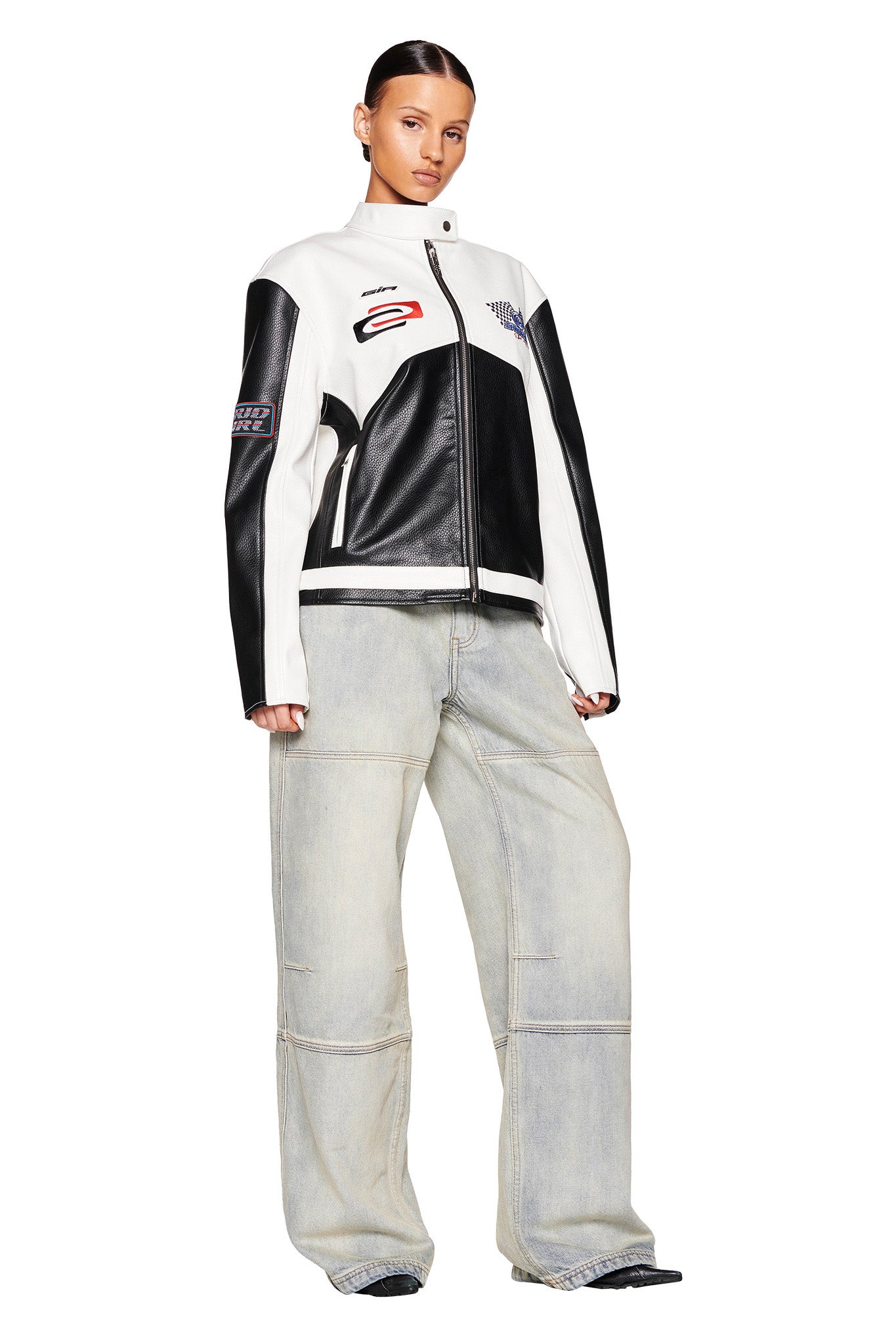 A person with slicked-back hair is wearing a black and white I.AM.GIA moto-style jacket adorned with various patches, paired with I.AM.GIA's MISTY JEAN - BLUE : VINTAGE WASH and black shoes. They stand with their hands by their sides, looking towards the camera against a white background.