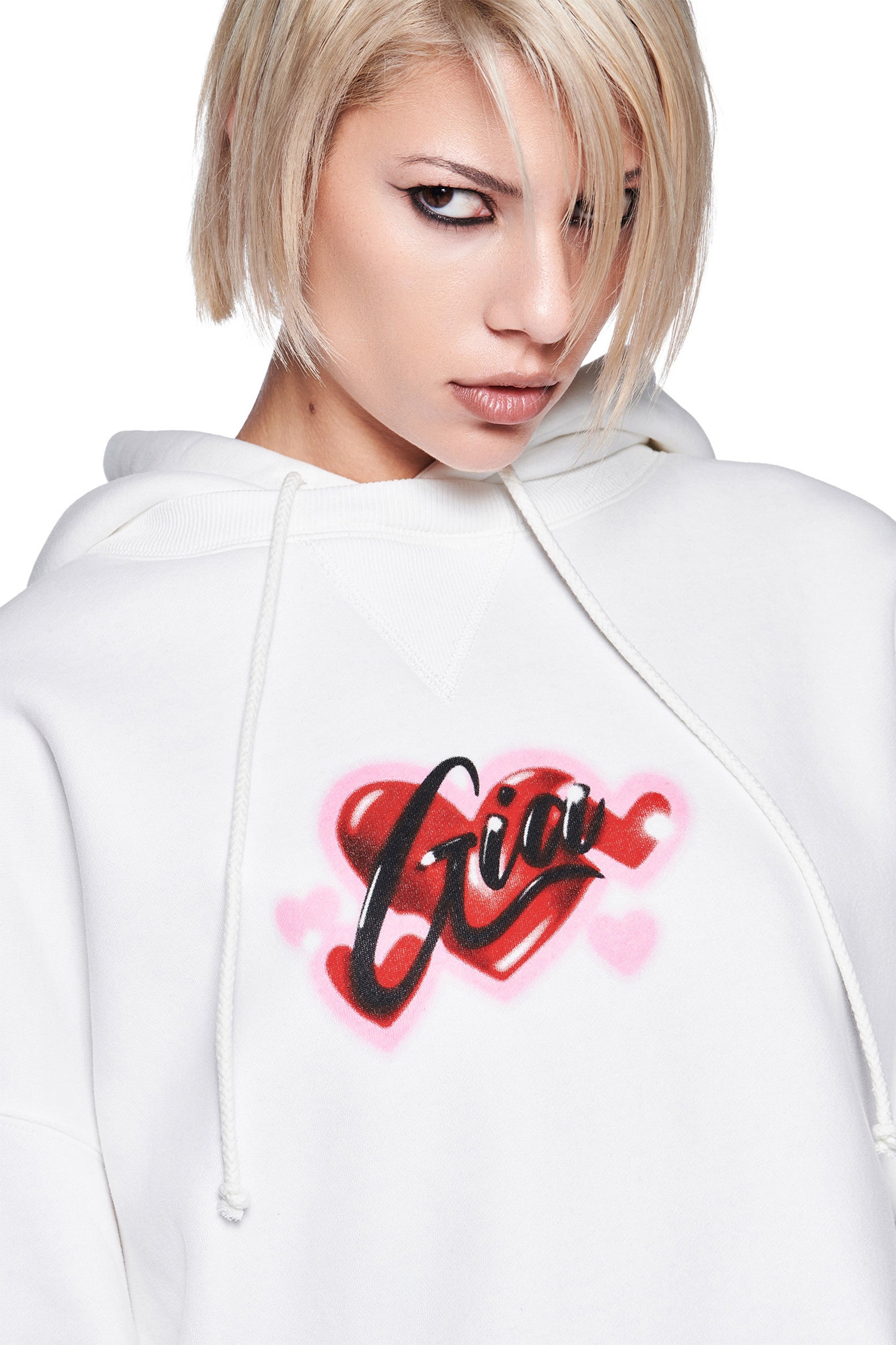 A person with short blonde hair and bold eye makeup wears an oversized white DORIAN HOODIE by I.AM.GIA, featuring a red heart and "Gia" in airbrush art. The background is plain white.