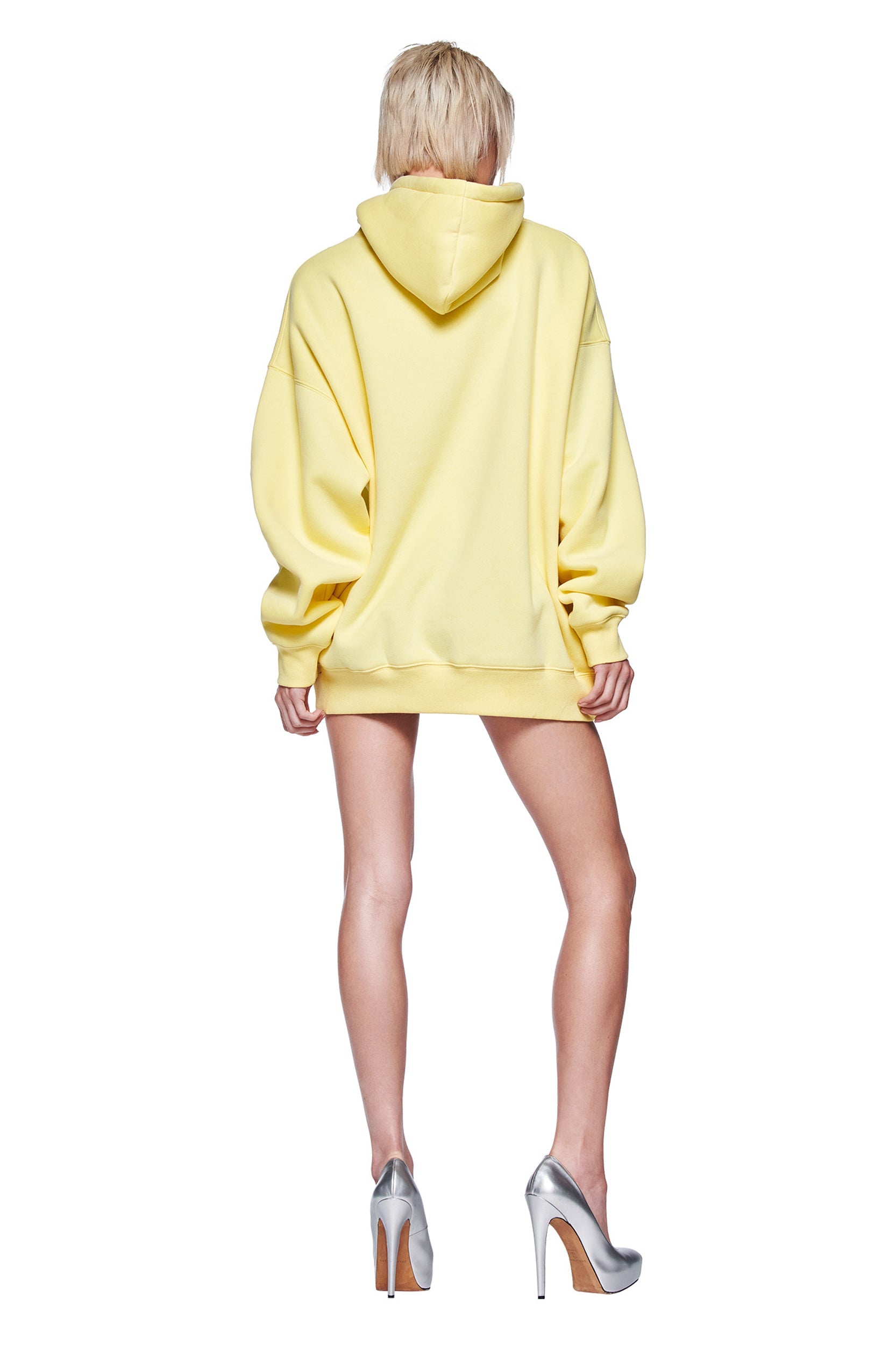 A person with short blond hair faces away, wearing the oversized I.AM.GIA DORIAN HOODIE in yellow and metallic silver heels against a white background—ideal for fans of bold, custom I.AM.GIA style.