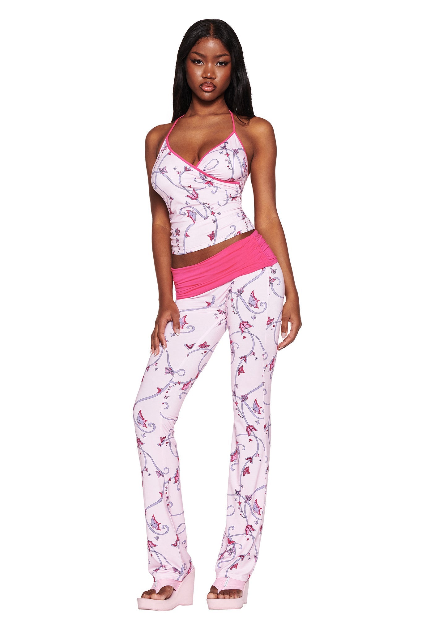 A woman poses confidently in the JANNIS PANT - PINK : BUTTERFLY RIBBON by I.AM.GIA, featuring a floral-patterned halter top and mid-rise pants with a bright pink waistband. Her long dark hair is styled down against a plain white background.