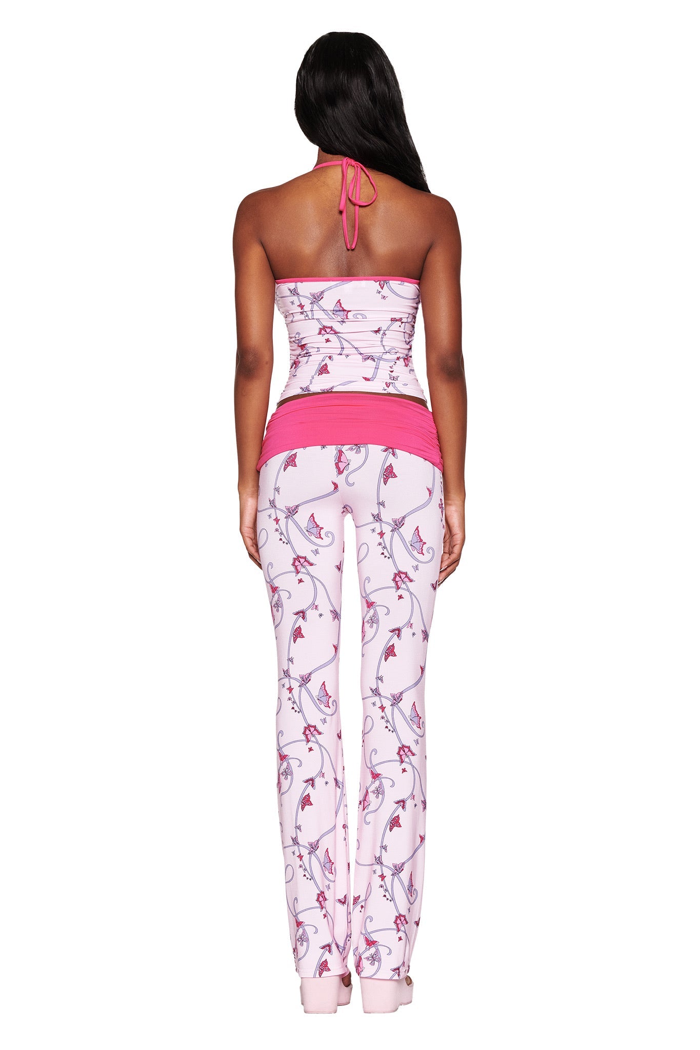 A person with long hair faces away, wearing the I.AM.GIA JANNIS PANT - PINK : BUTTERFLY RIBBON. The outfit features a pink halter top, floral-patterned leggings with butterfly ribbon print, a pink waistband, and abstract floral designs against a plain white background.
