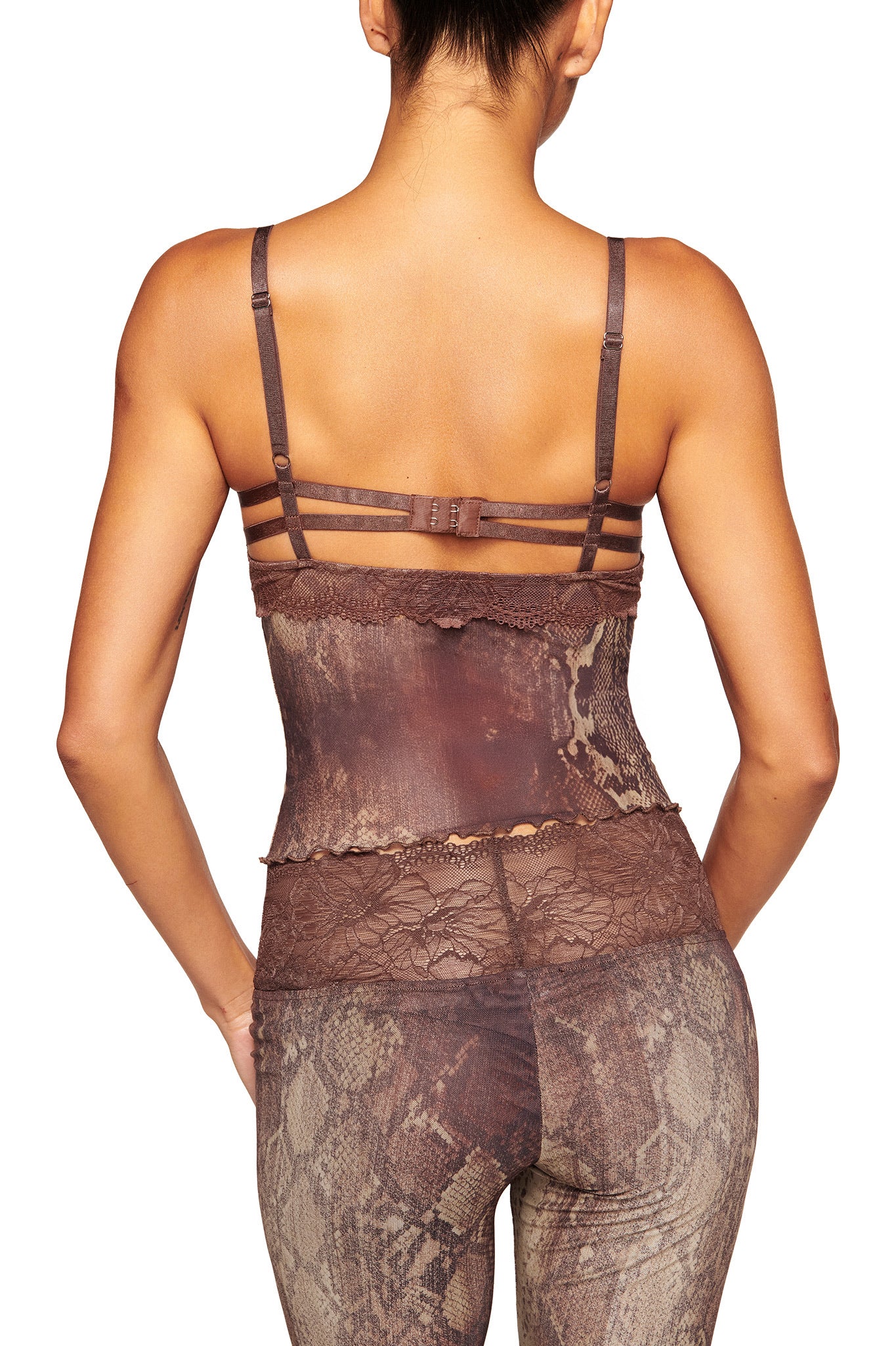 A woman with her hair tied up is seen from the back wearing I.AM.GIA's brown, strappy lace camisole paired with SILO PANT - SNAKE high-waisted snake print pants. Her arms rest relaxed at her sides.