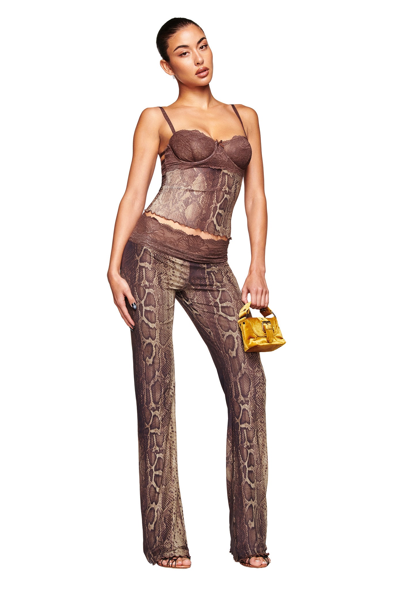 A woman models the I.AM.GIA SILO PANT - SNAKE, a sheer brown snake print pants set with matching camisole, accessorized with a gold handbag and strappy heels. She poses against a white background with her hair pulled back.