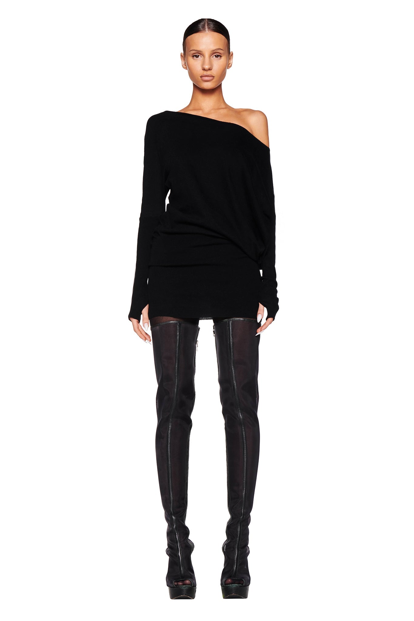 A person with slicked-back hair is wearing the black GISELLE MINI DRESS by I.AM.GIA and black over-the-knee boots. They are standing against a white background, gazing directly at the camera.
