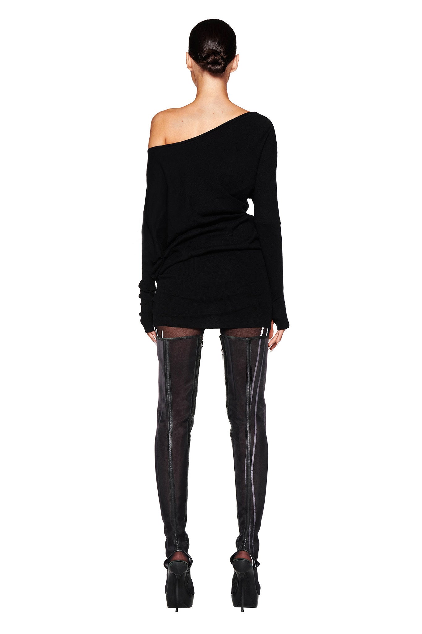 A person is standing facing away from the camera, wearing an I.AM.GIA GISELLE MINI DRESS - BLACK paired with thigh-high black boots featuring back zipper details. Their hair is styled in a sleek bun. The image is set against a white background.