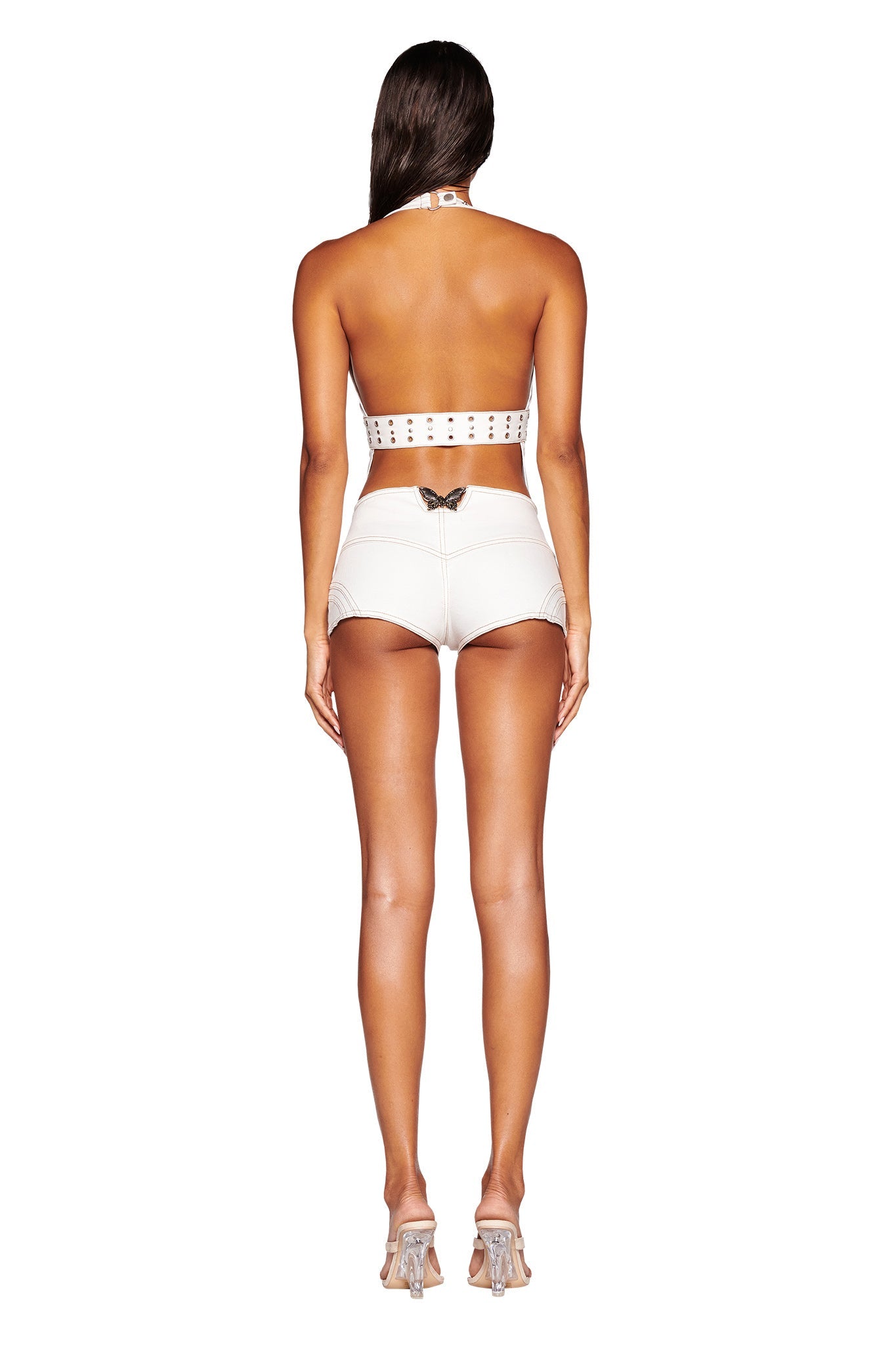 A person is standing with their back facing the camera, donning a white halter top and SEQUOIA SHORT - WHITE : DENIM by I.AM.GIA, paired with clear heeled sandals. The low-rise waist shorts feature a small decorative butterfly detail at the waistband. The individual has long brown hair.