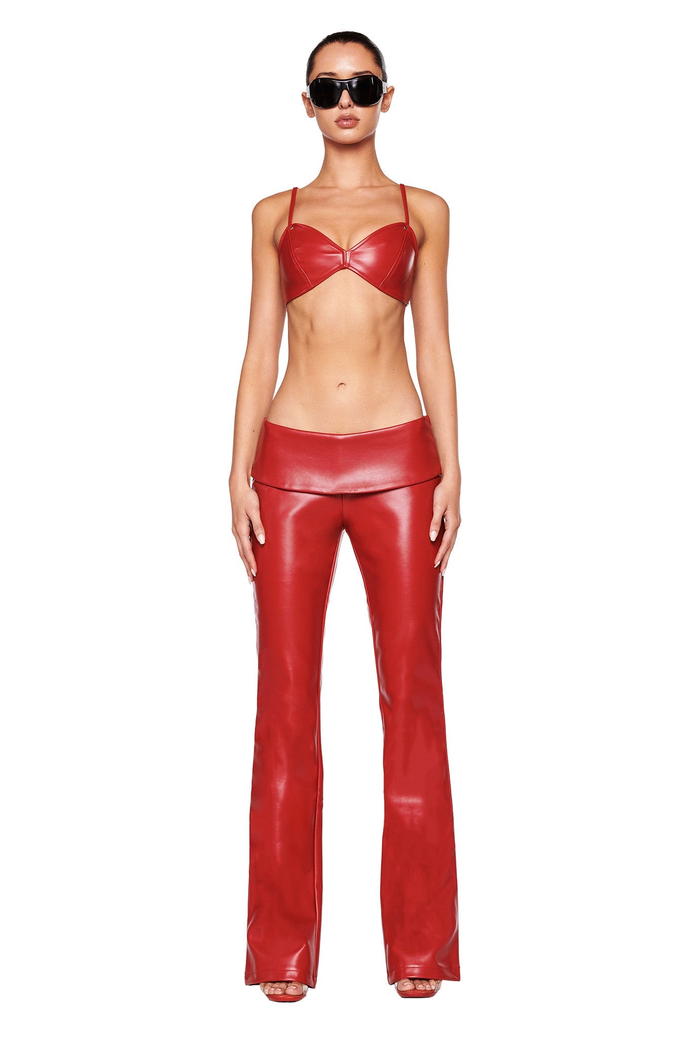 A woman stands facing the camera wearing an I.AM.GIA red leather two-piece outfit consisting of a crop top and AMALTHEA PANT 2.0 - RED. She is also wearing black sunglasses and red heeled sandals. The background is plain white.
