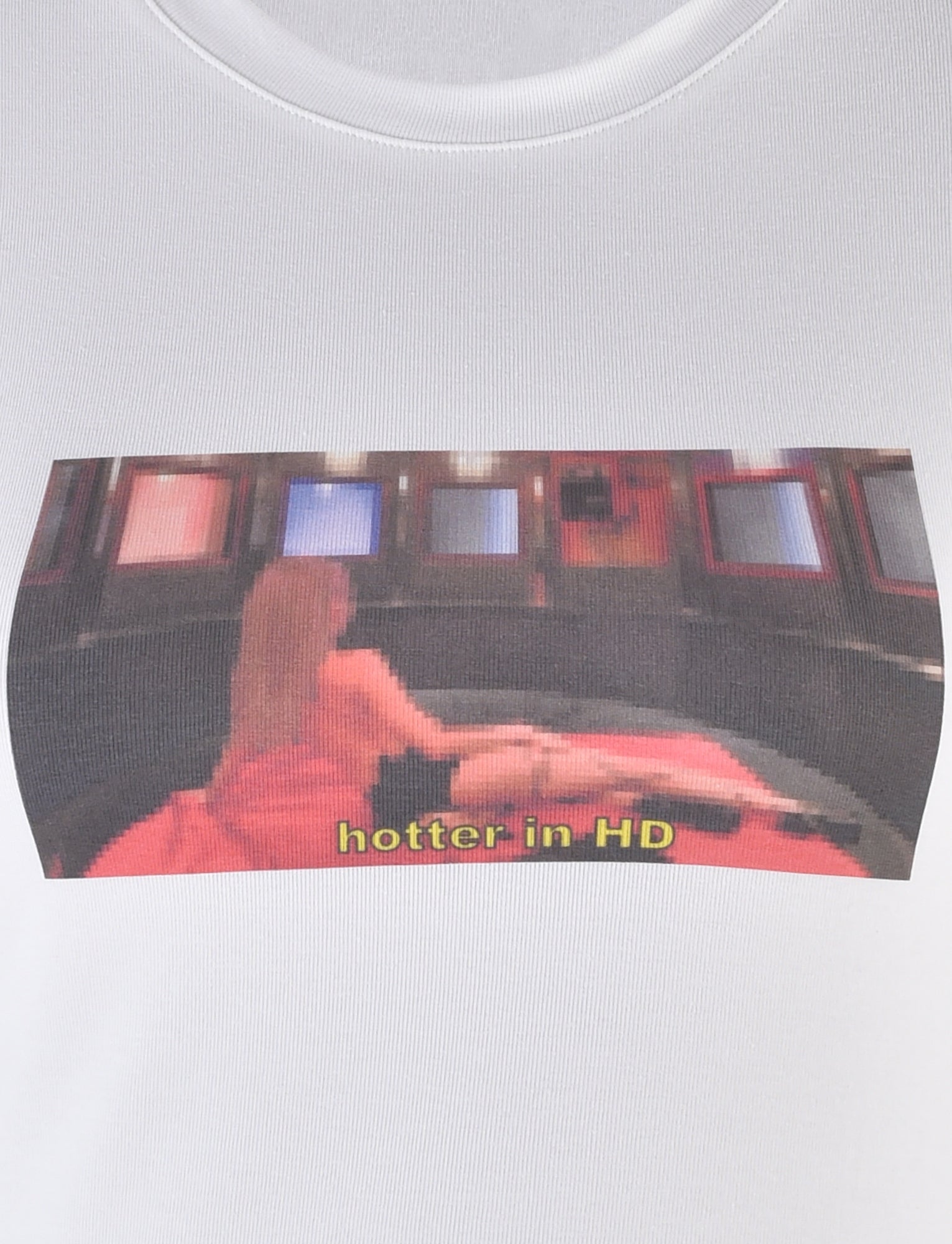 The ODEZIA TOP - WHITE by I.AM.GIA features a white fabric with a graphic of a pixelated person sitting on a circular couch surrounded by multiple television screens. The phrase "hotter in HD" is prominently displayed at the bottom, capturing the edgy style that defines the I.AM.GIA brand.