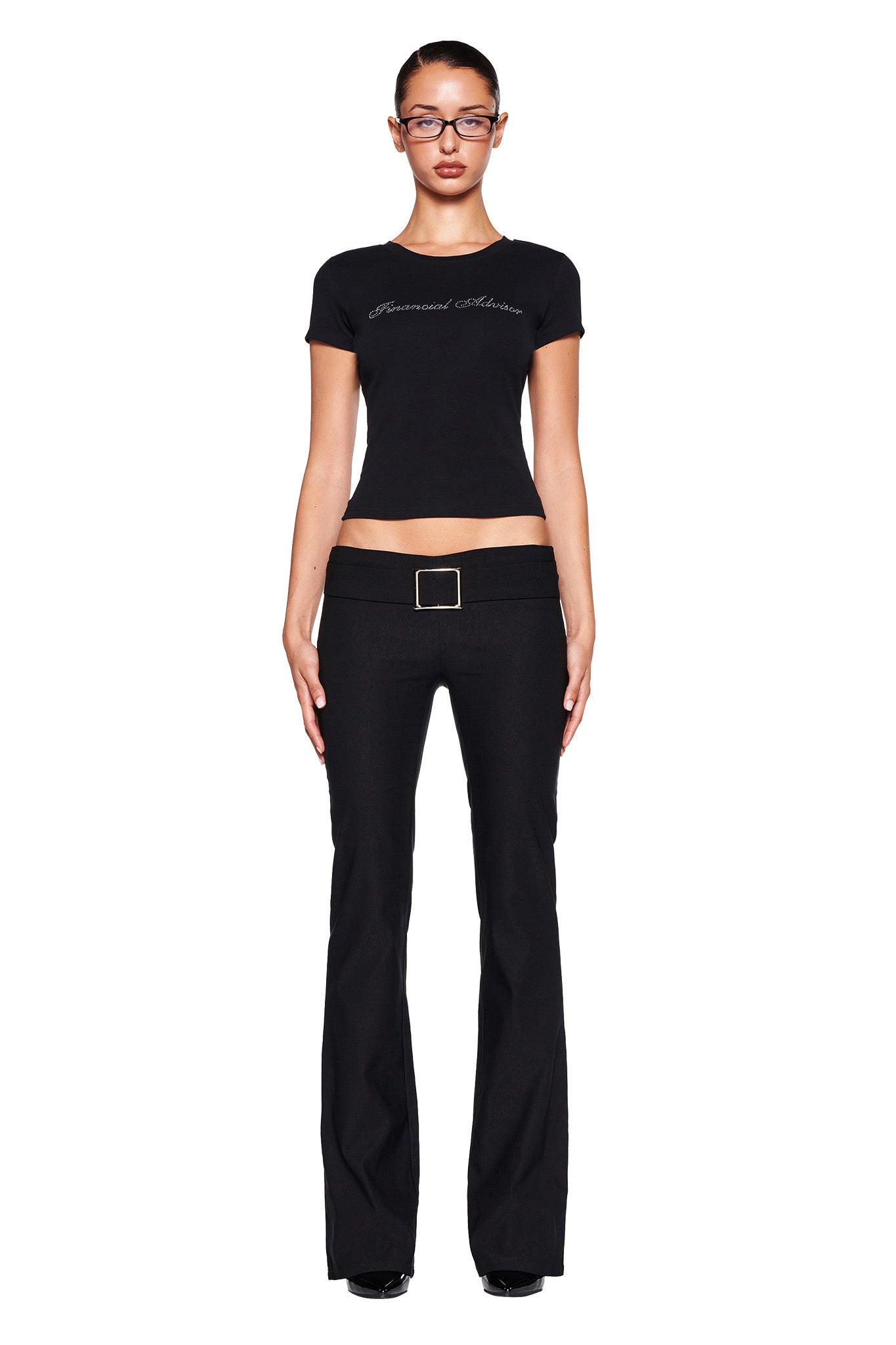 A person stands upright against a white background, wearing the ODEZIA TOP - BLACK:DIAMONTE from I.AM.GIA, which features a custom diamante print across the chest. They pair it with black flared pants and a belt with a rectangular buckle. The soft jersey fabric of the top fits perfectly while their arms rest at their sides, and they are wearing glasses.